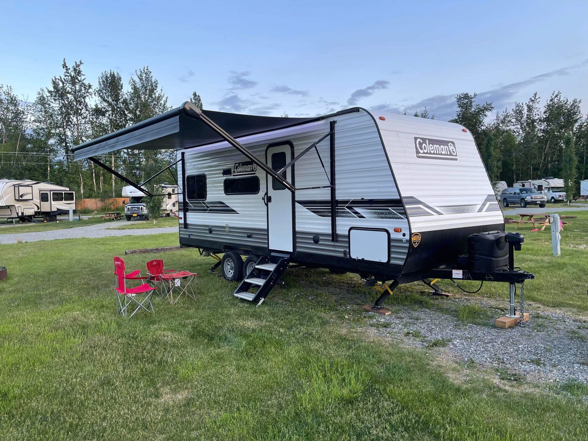 A white Coleman travel trailer, possibly an RV rental, is parked on a grassy site with a deployed awning & camping chairs. Ideal for outdoor adventures.. Dutchmen Coleman lantern LT 2022