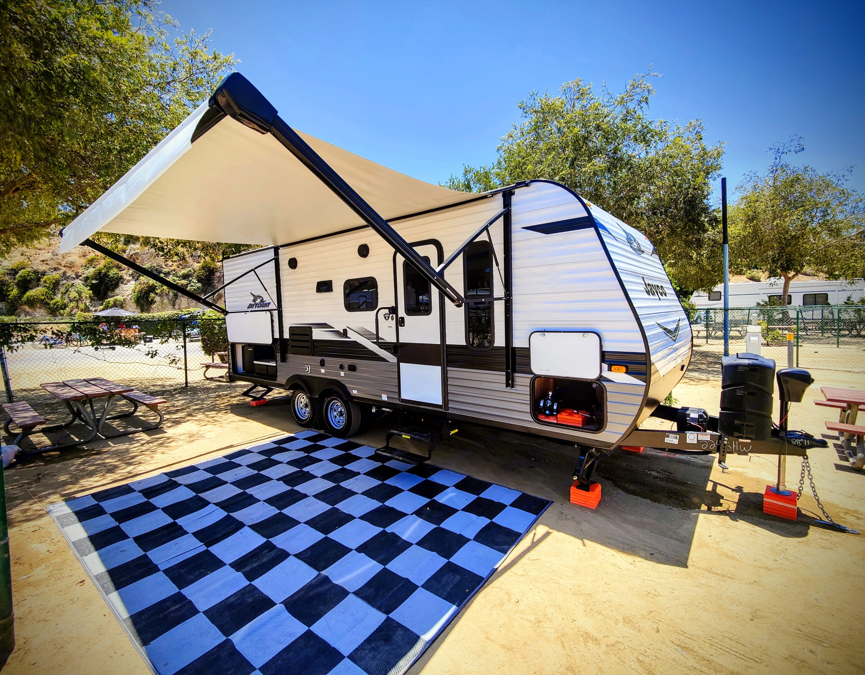 Bright travel trailer RV rental at a sunny campsite, awning extended over a blue checkered mat. Perfect for outdoor adventures.. Jayco Jay Flight SLX 2022