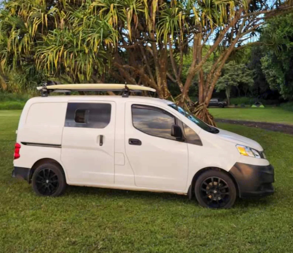 White camper van with surfboard on roof, parked on grass at a. Nissan NV200 2018