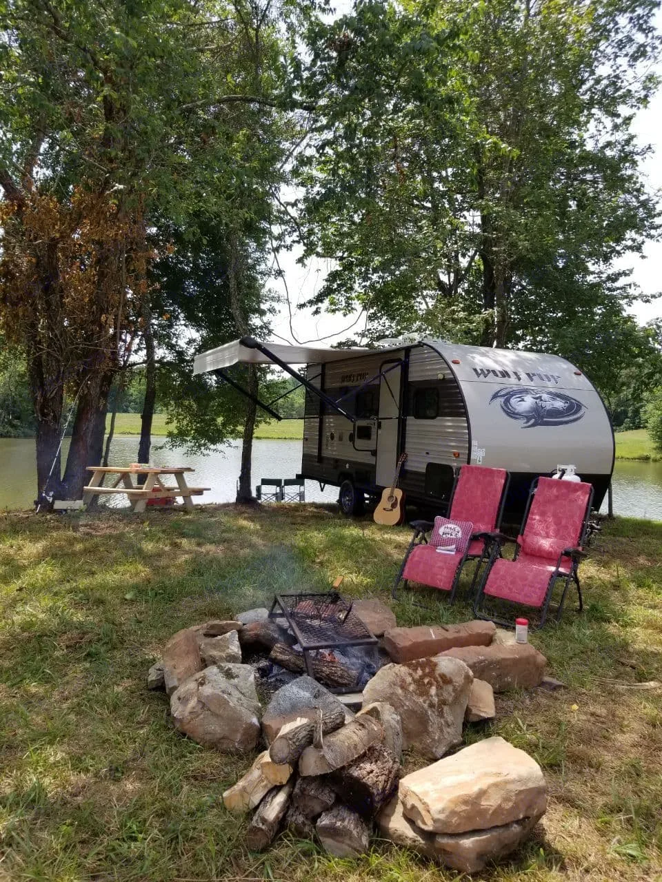 Serene lakeside RV rental setup with a cozy campfire, red chairs, and a picnic table, perfect for outdoor camping.. Forest River Cherokee Wolf Pup 2018