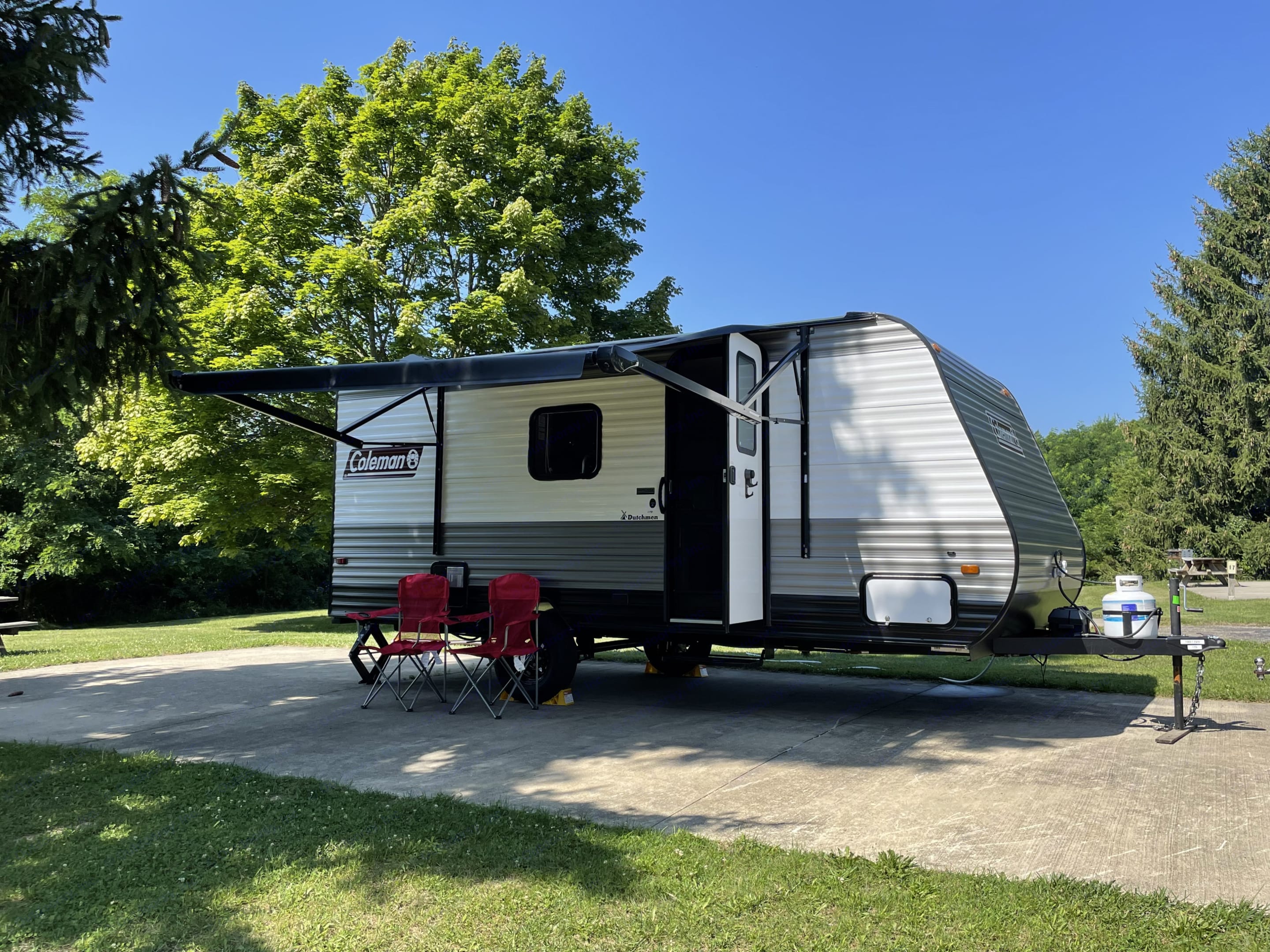 A modern travel trailer RV rental, grey and white, under a blue sky at a sunny campsite with its awning extended and red chairs.. Dutchmen Coleman Lantern LT 2022