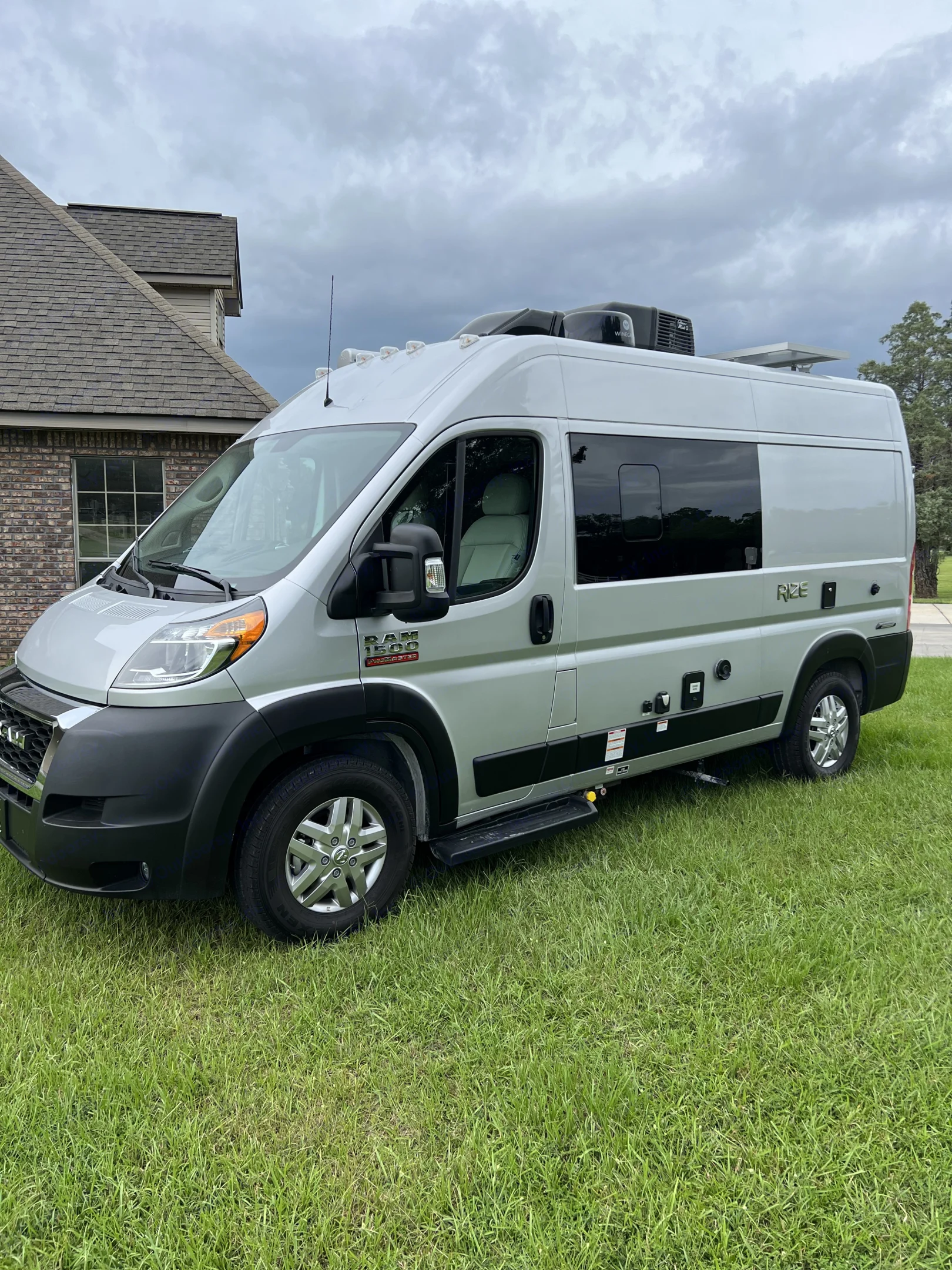 Silver Ram ProMaster camper van parked on green grass, ready as an RV rental. Visible features include a deployed step and roof units.. Thor Rize 2022
