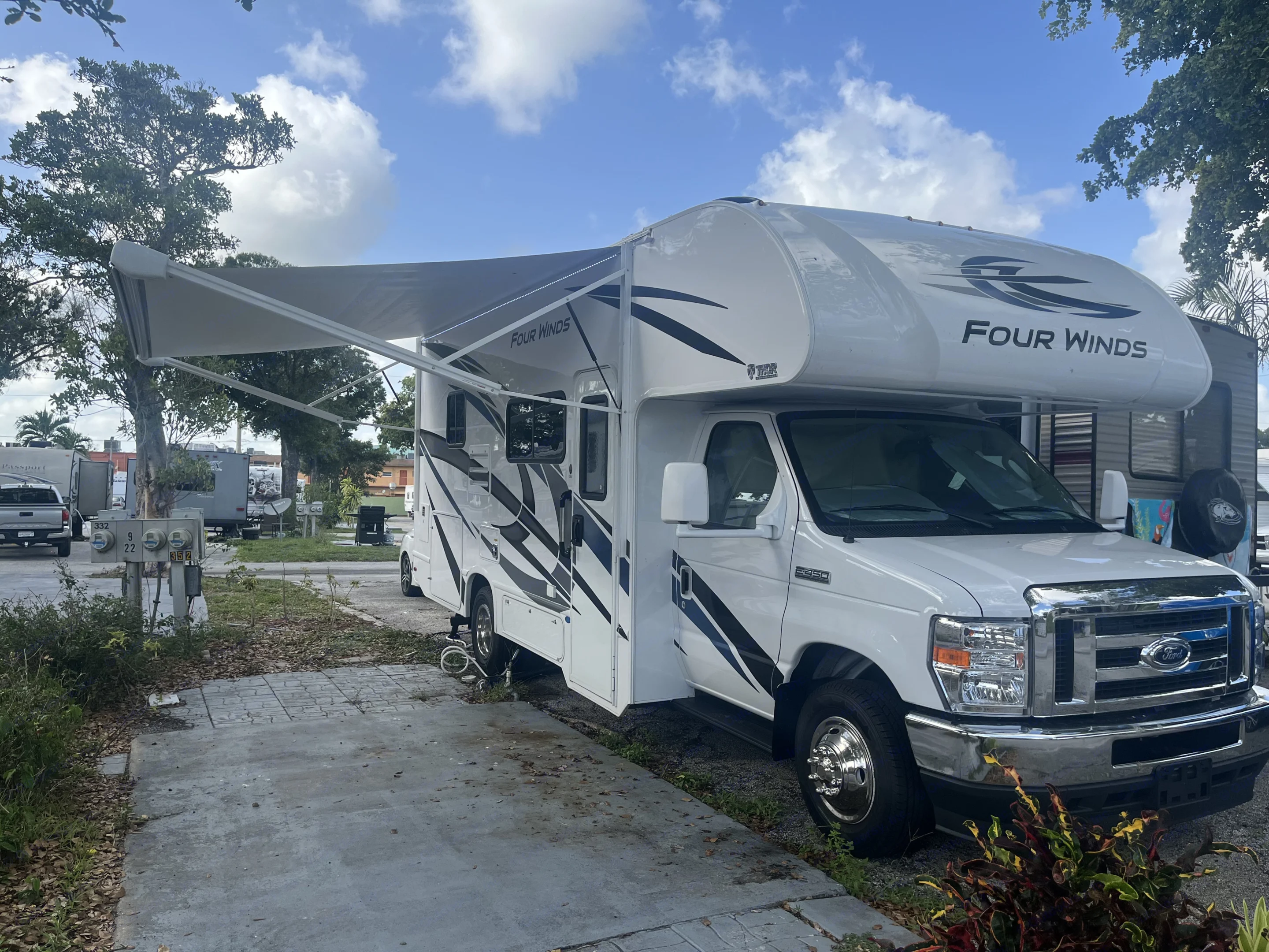 White Four Winds RV rental parked at a sunny campsite with its awning extended. Utility hookups visible.. Thor Four Winds 2023