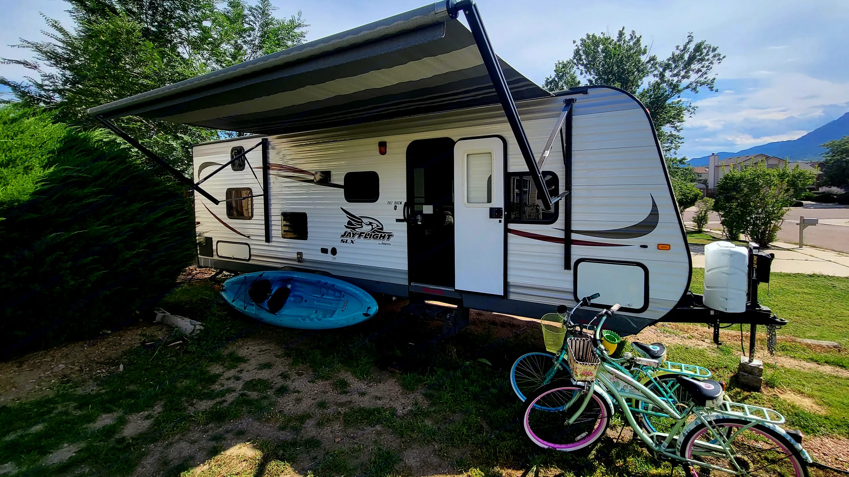 An RV rental, complete with an extended awning, a blue kayak, and two bicycles, is parked in a grassy yard near mountains.. Jayco Jay Flight SLX 2015