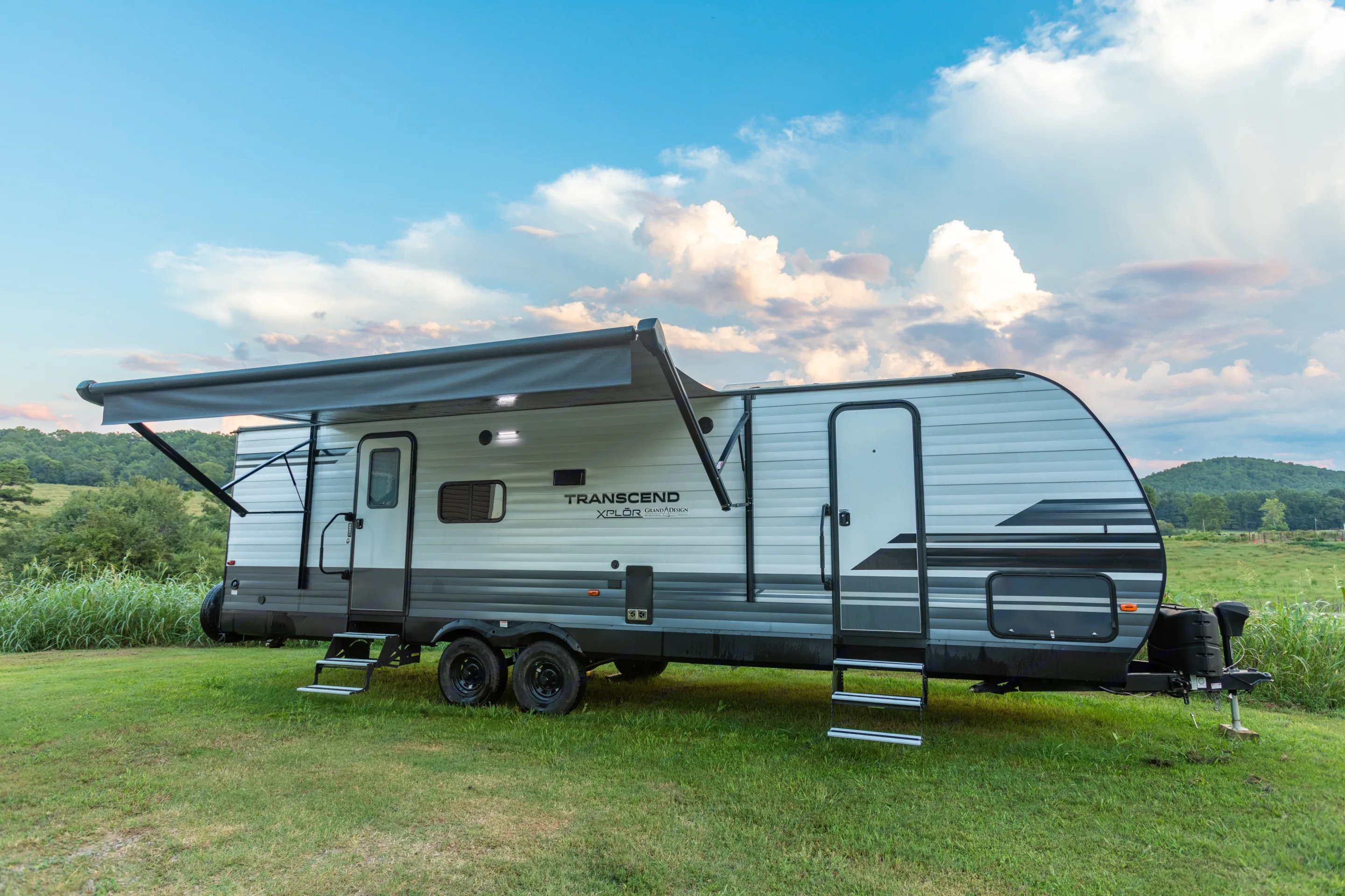 An RV rental, a grey and white travel trailer with an extended awning, sits on green grass under a partly cloudy sky.. Grand Design Transcend Xplor 2021