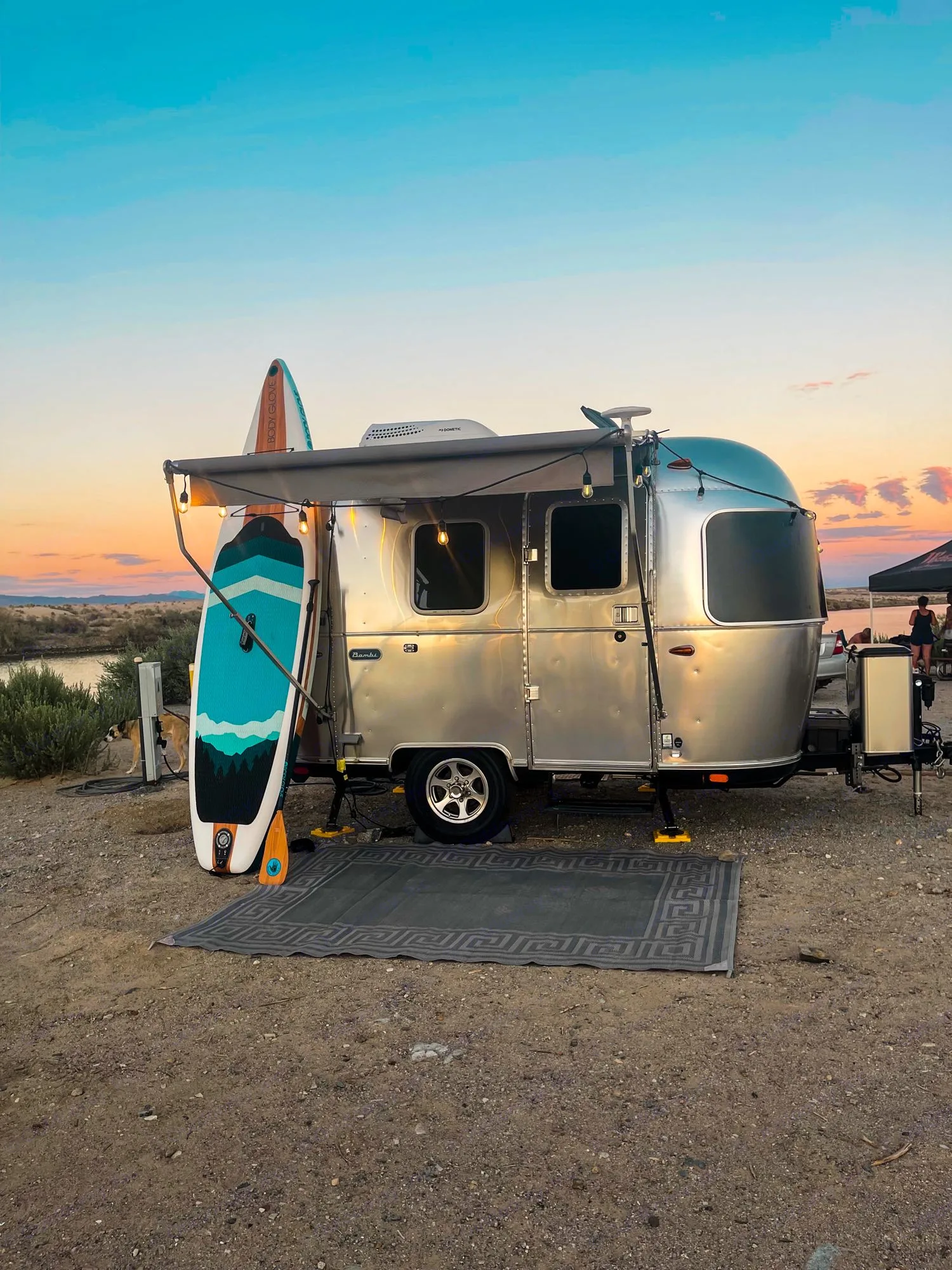 An Airstream RV rental parked by the water at sunset with paddleboards. A cozy camping scene ready for adventure.. Airstream Bambi 2021