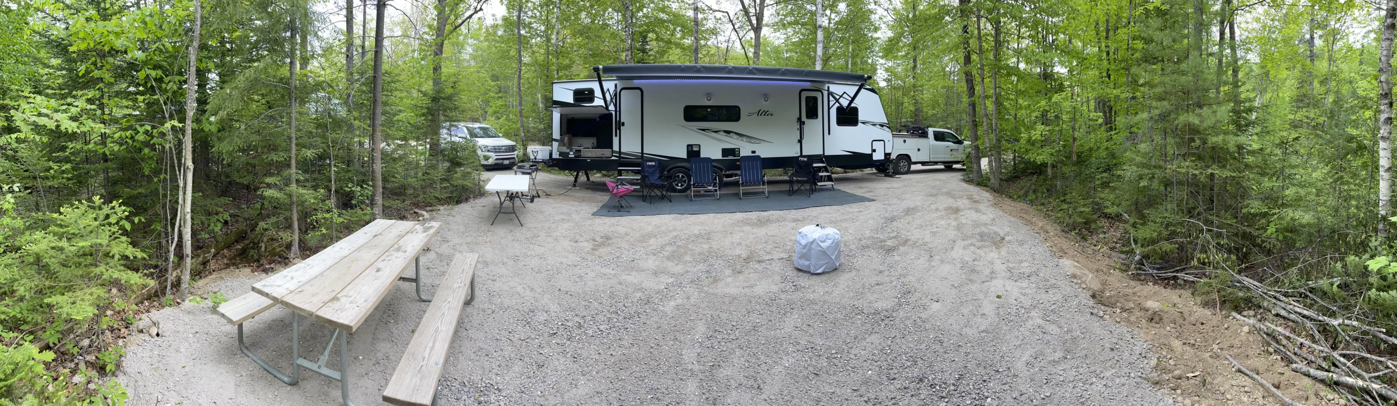 Scenic RV rental campsite setup: white travel trailer, outdoor kitchen, chairs, picnic table, and truck surrounded by trees.. East to West Alta 2022