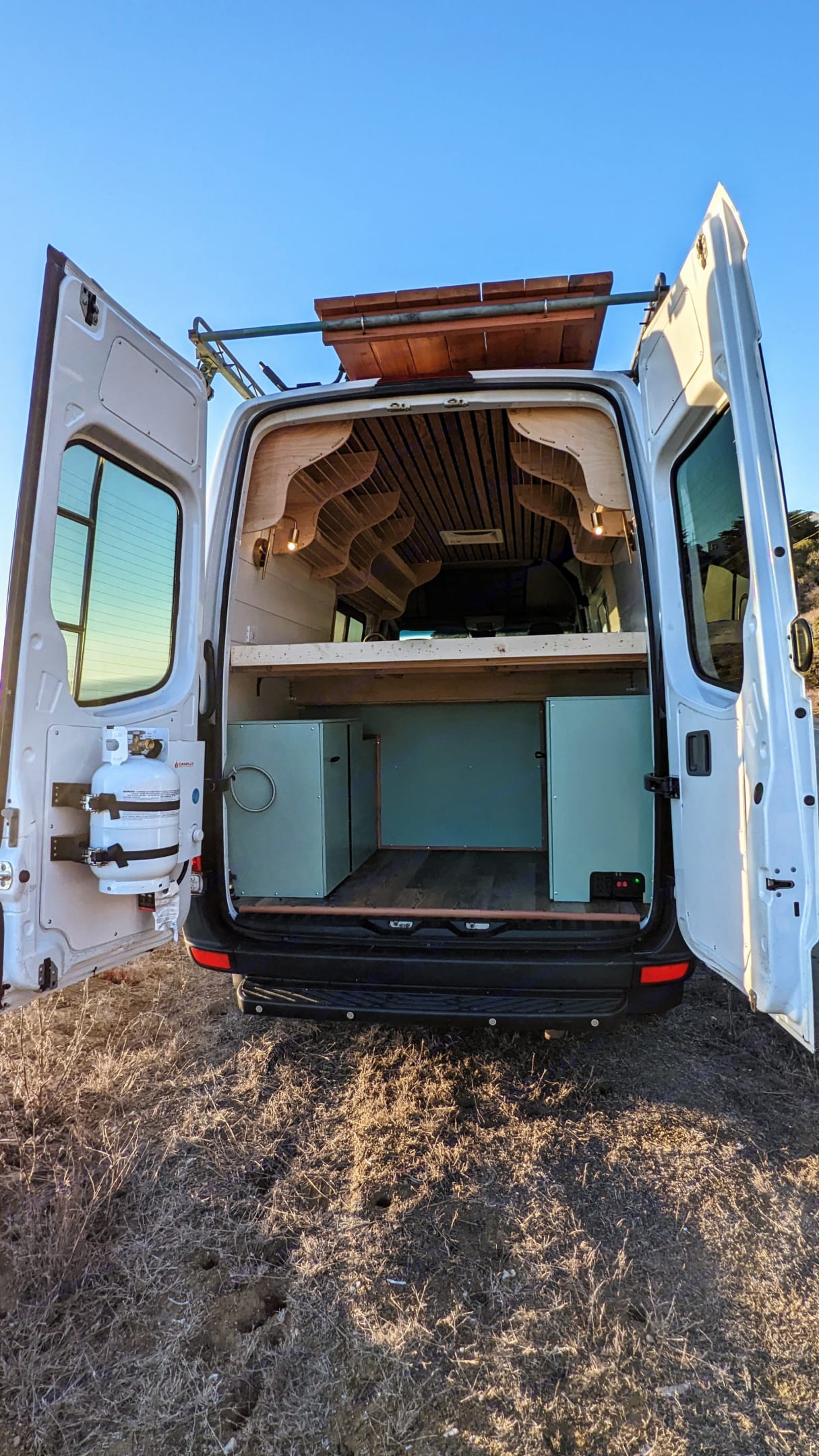 An RV rental camper van with open rear doors reveals a custom wood interior, bed, and storage. Ideal for road trips.. Mercedes-Benz Sprinter 2014