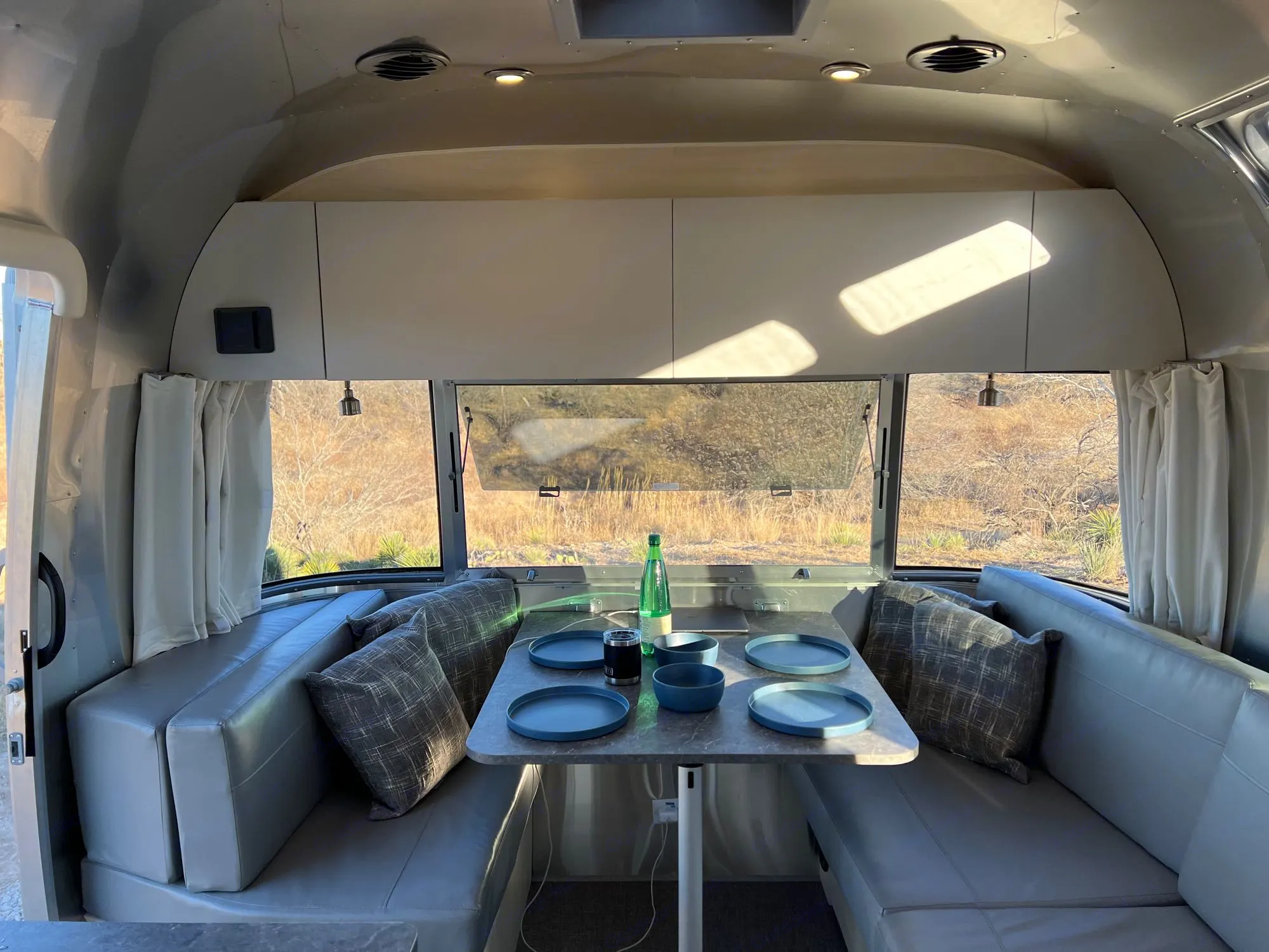 Inside an RV rental: a modern dining area with a table set for four, surrounded by comfortable benches and large windows showcasing nature.. Airstream Flying Cloud 2023