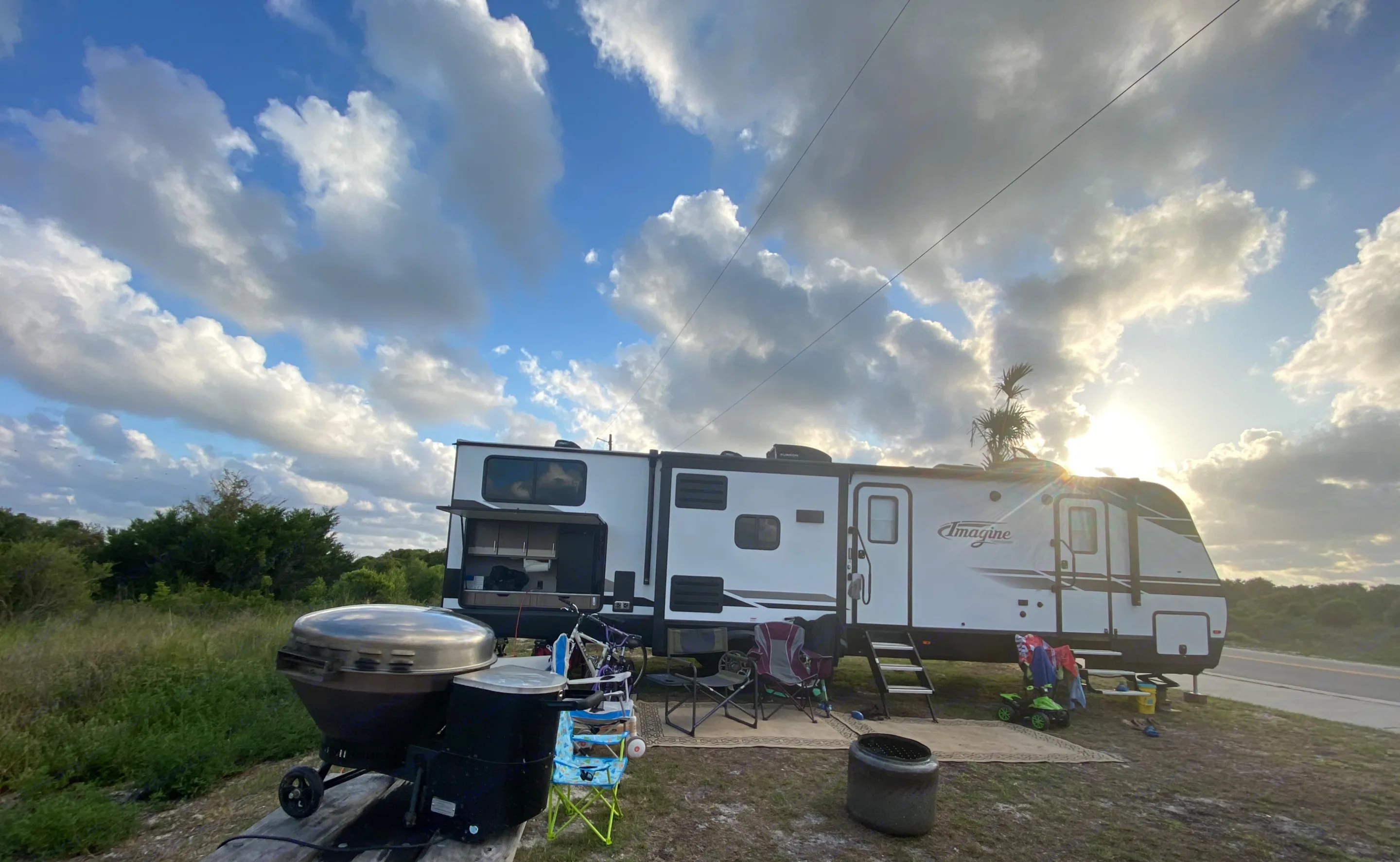Scenic RV rental campsite with an outdoor kitchen, grill, and chairs, bathed in sunlight under a cloudy sky.. Grand Design Imagine 2022