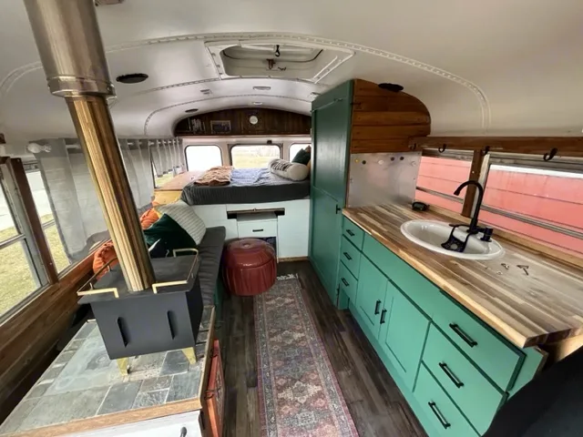 Converted bus RV rental showcasing a modern kitchen with teal cabinets, wood stove, and rear bed setup.. GMC Savana 2002