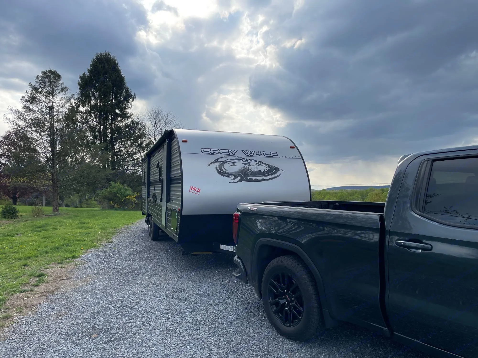 A dark truck is hitched to a silver Grey Wolf travel trailer (a common RV rental type) on a gravel drive. Cloudy sky.. Forest River Cherokee Grey Wolf 2019