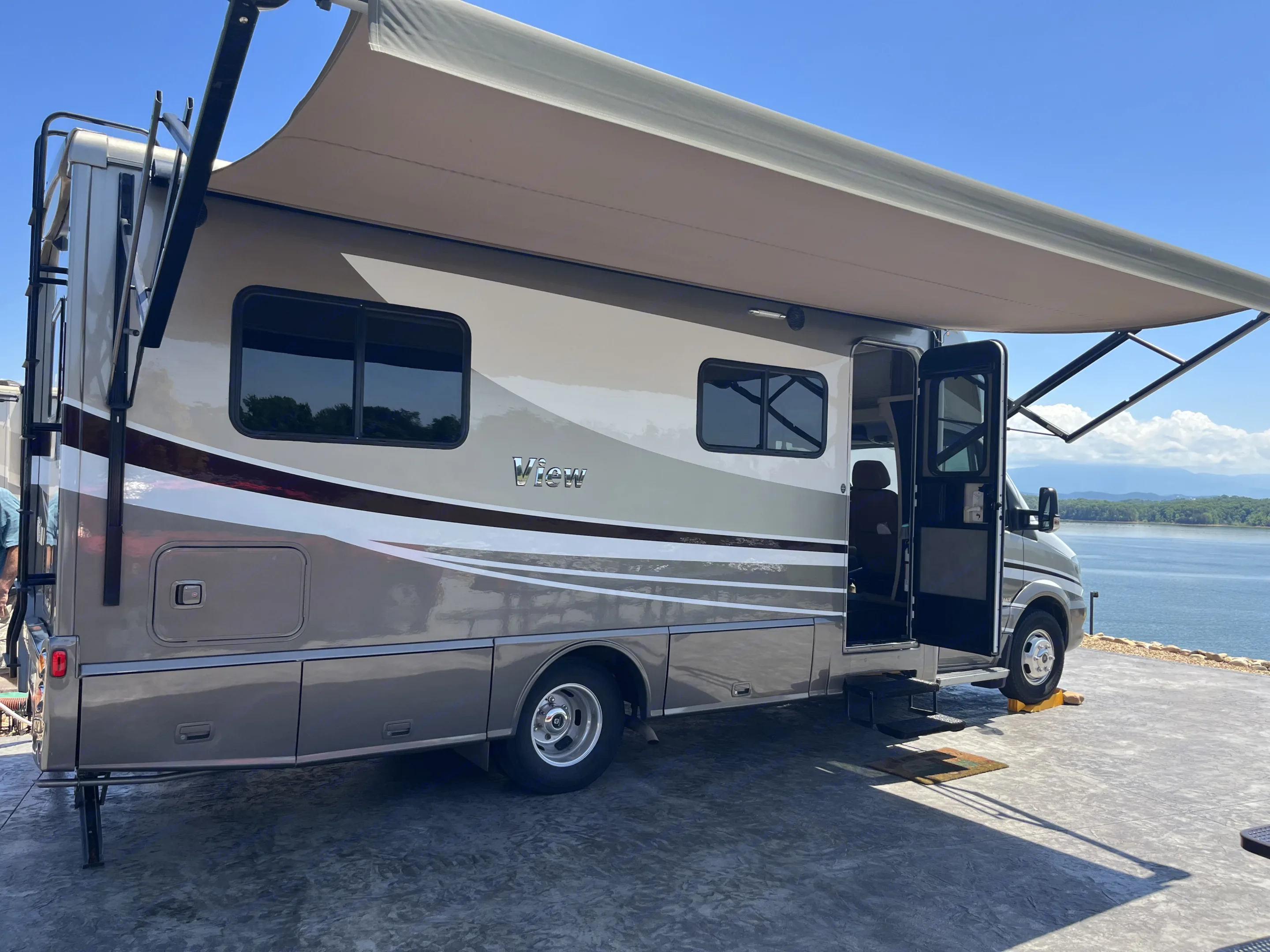 This spacious rv rental, a "View" model, is parked by a scenic lake with its awning extended for shade.. Winnebago View 2018