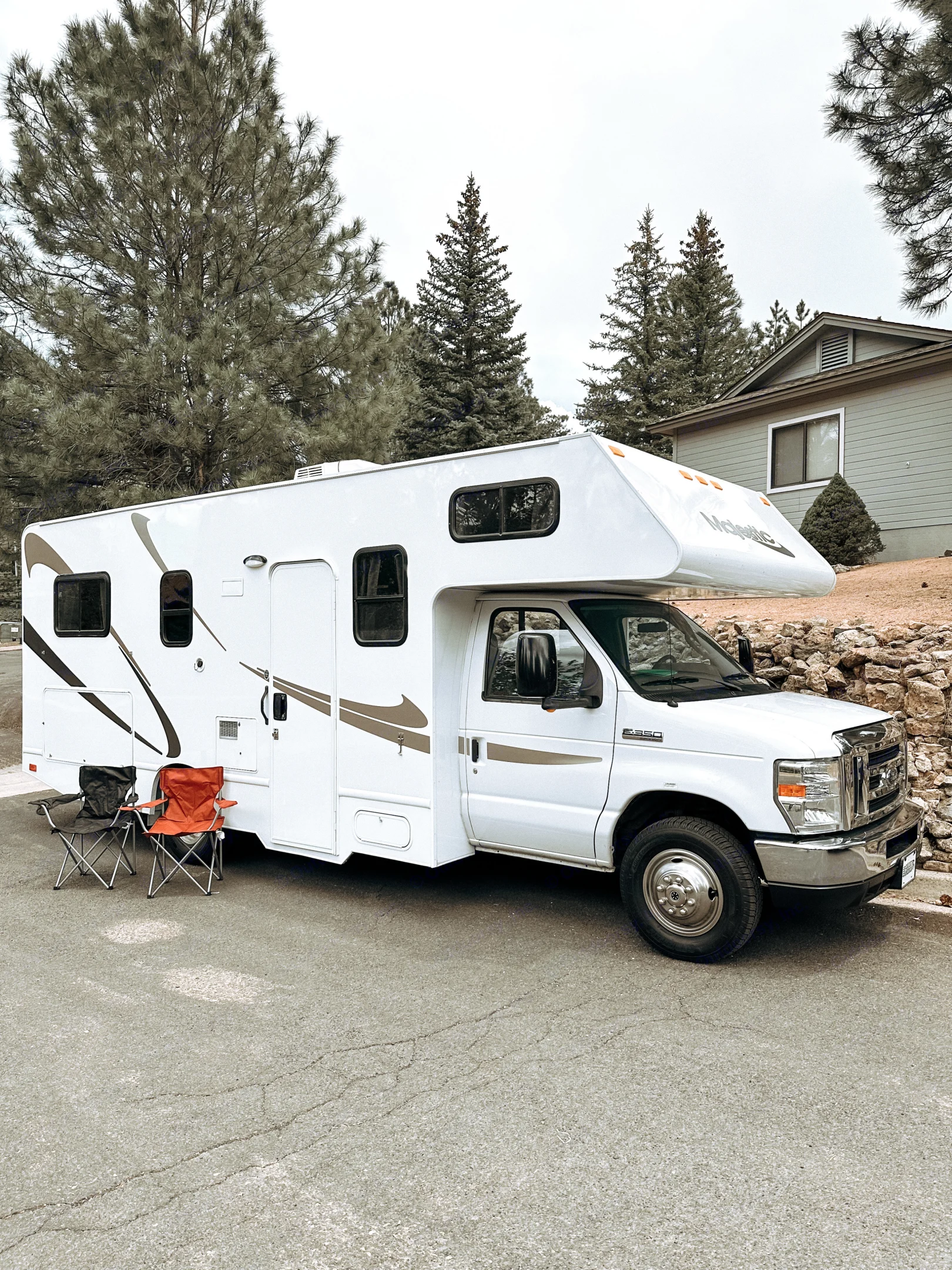 This white RV rental, a Majestic Class C, is parked on an asphalt road with chairs ready for camping. Tall trees & a house visible.. Thor Majestic 2019