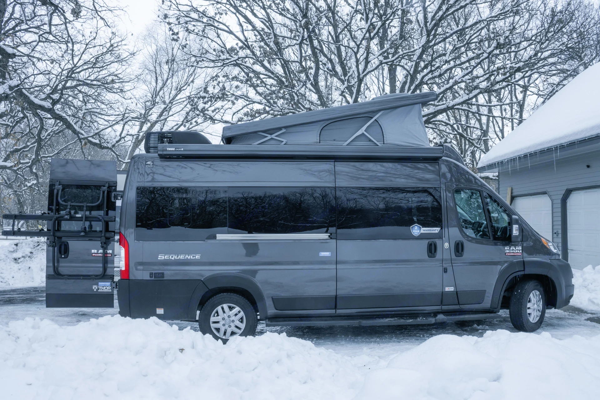 Grey camper van RV rental with pop-top roof and rear rack, parked in a snow-covered yard. Ready for winter travel!. Thor Sequence 2022