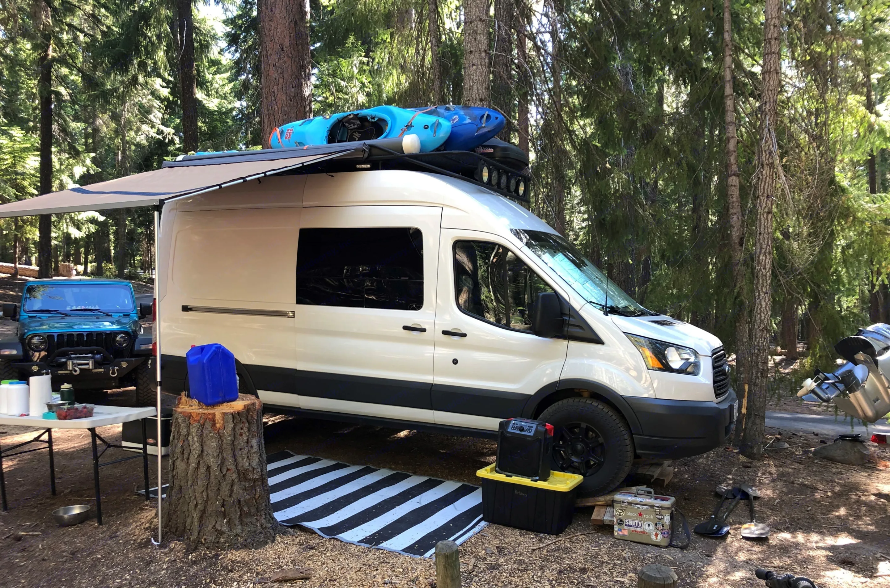 White camper van with awning & kayaks parked in a forest campsite next to a blue Jeep. Ideal for an RV rental adventure!. Ford Transit 2016