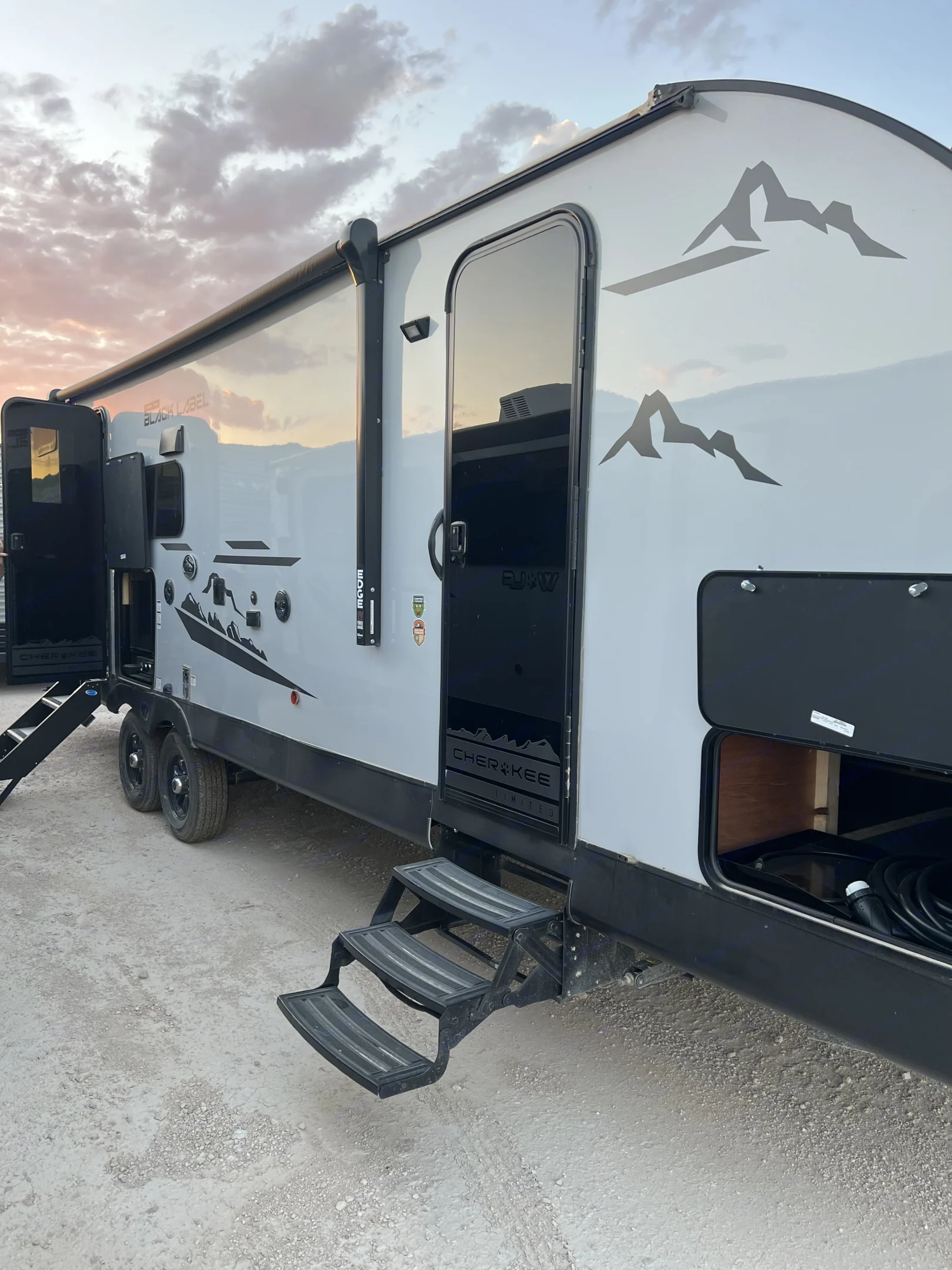 A modern gray Cherokee **rv rental** with mountain decals, open doors, and steps, parked on gravel under a sunset sky.. Forest River Cherokee Grey Wolf 2023
