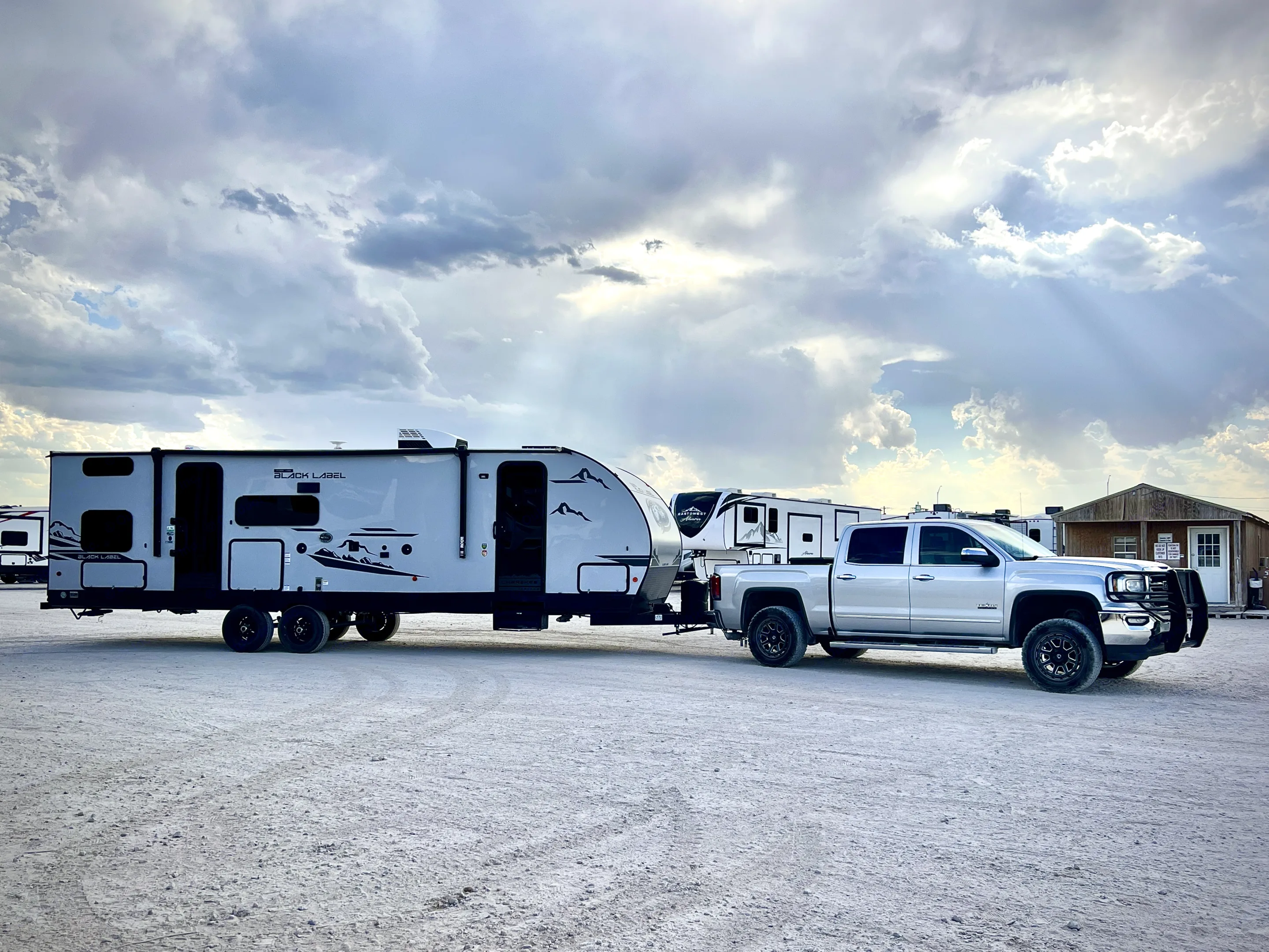 A pickup truck towing a gray 'Black Label' RV travel trailer is parked on a sandy lot. Clouds with sunbeams highlight this potential RV rental setup.. Forest River Cherokee Grey Wolf 2023