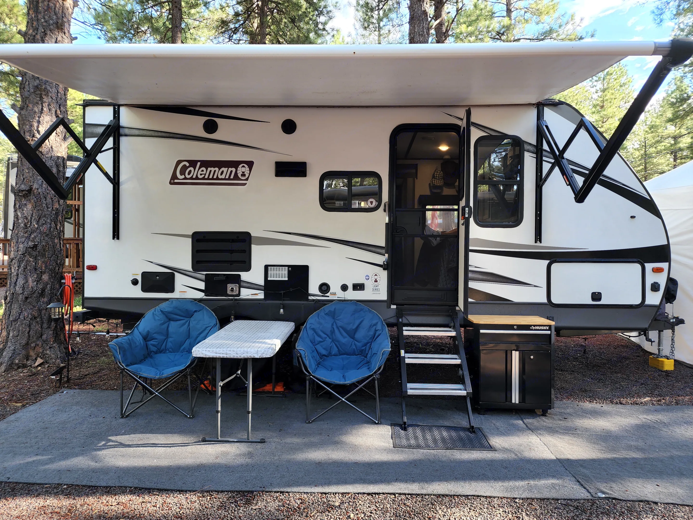An inviting rv rental campsite: a white Coleman travel trailer with its awning extended, flanked by comfy blue chairs and a table.. Dutchmen Coleman Light 2020