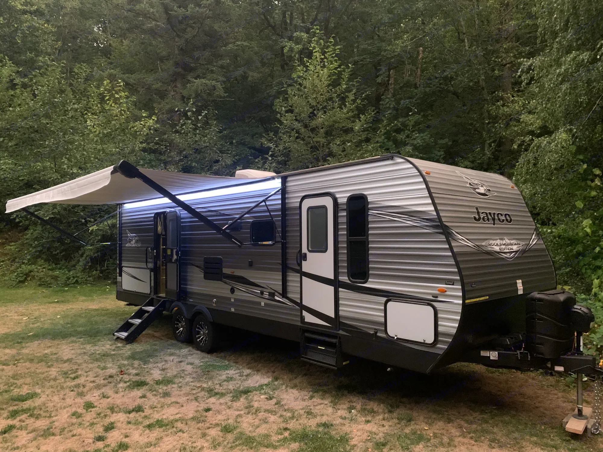 This Jayco travel trailer, likely an RV rental, is set up for camping at dusk, with its awning lights. Jayco Jay Flight SLX Rocky Mountain Edition 2020