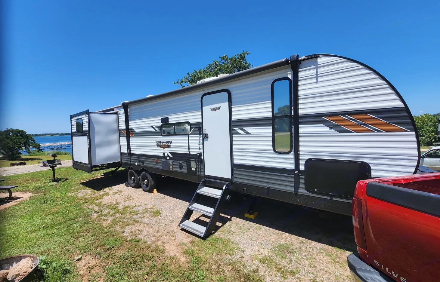 A large, modern travel trailer RV rental is parked at a sunny lakeside campsite with its slide-out extended, offering ample space.. Forest River Wildwood 2022