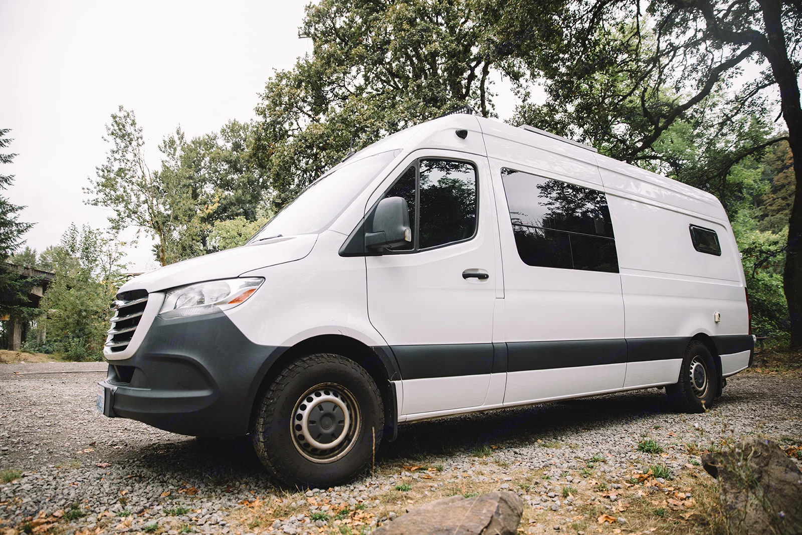 White camper van for RV rental parked on a gravel path, ready for outdoor adventures amidst lush trees.. Mercedes Sprinter 2019