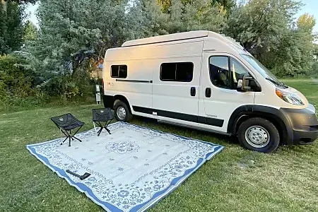White RV rental van parked on grass with an outdoor mat and chairs. Winnebago Solis 2023