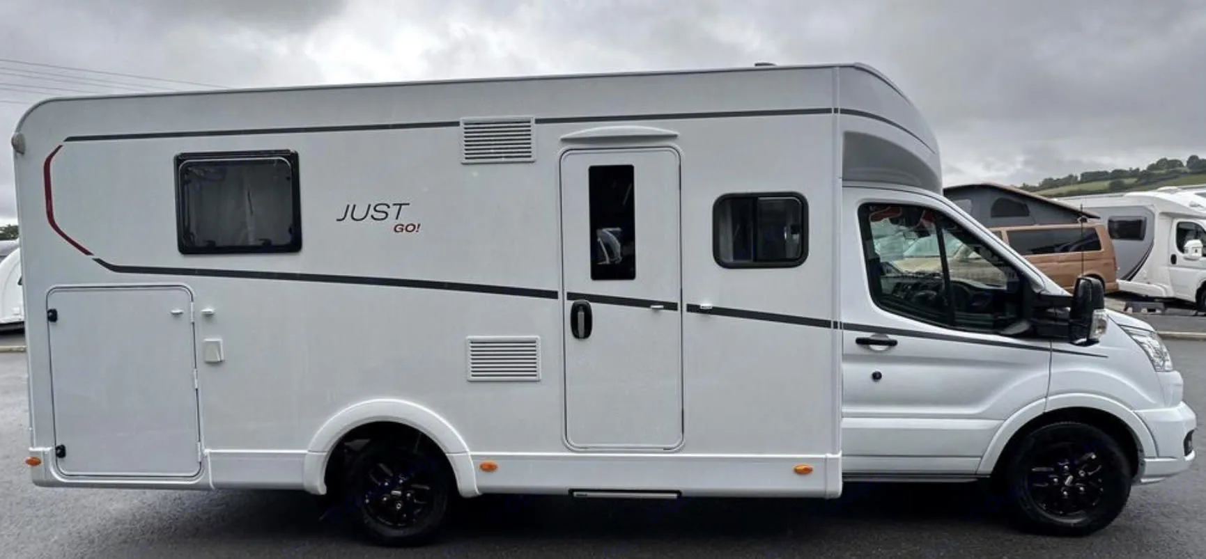 Side view of a white "JUST GO!" RV rental, a motorhome with black wheels, parked outdoors under a cloudy sky.. Dethleffs Just Go 2023