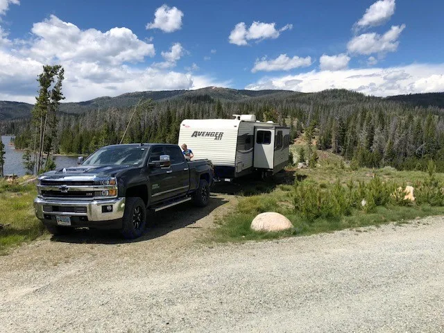 Chevy pickup towing an RV rental travel trailer at a scenic lakeside spot with forested mountains under blue skies.. Avenger ATI 2018
