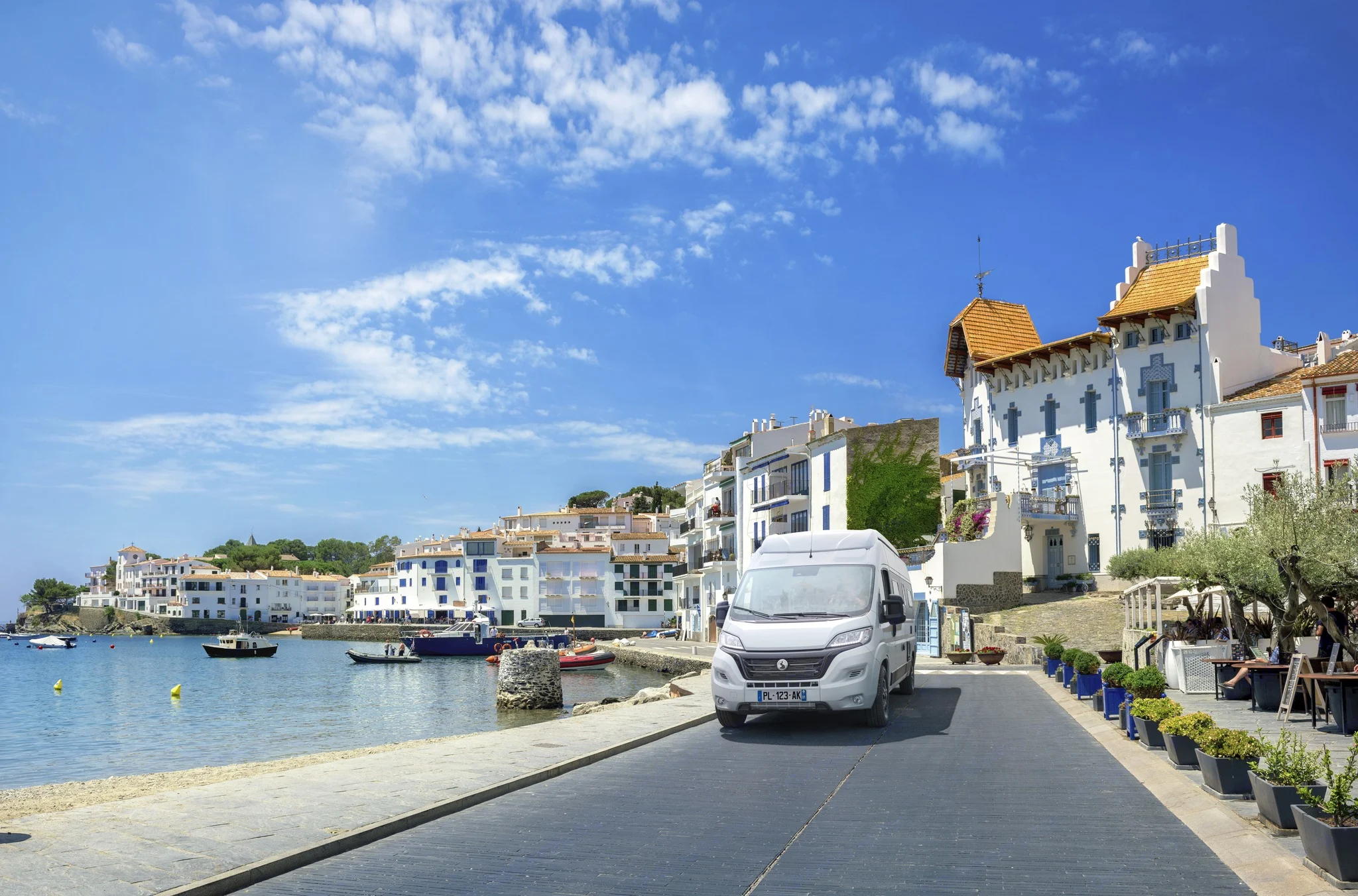 An RV rental drives along a sunny Mediterranean coast past white buildings and boats in a bay.. Bavaria K 600S 2024