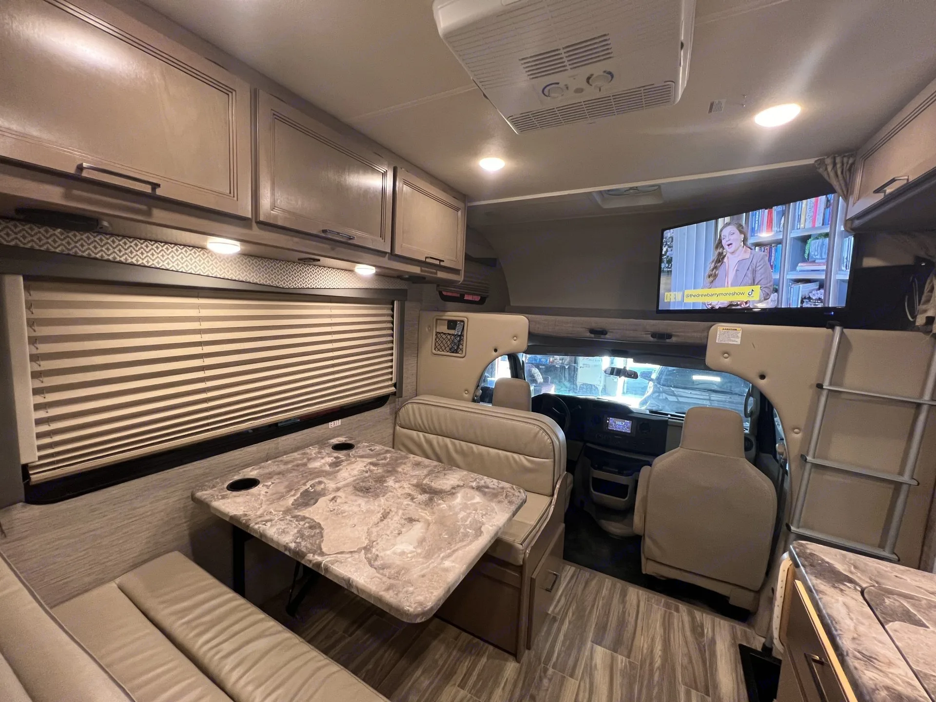 Bright RV rental interior features a dinette with benches & patterned table, overhead cabinets, TV, and cab view.. Thor Four Winds 2022