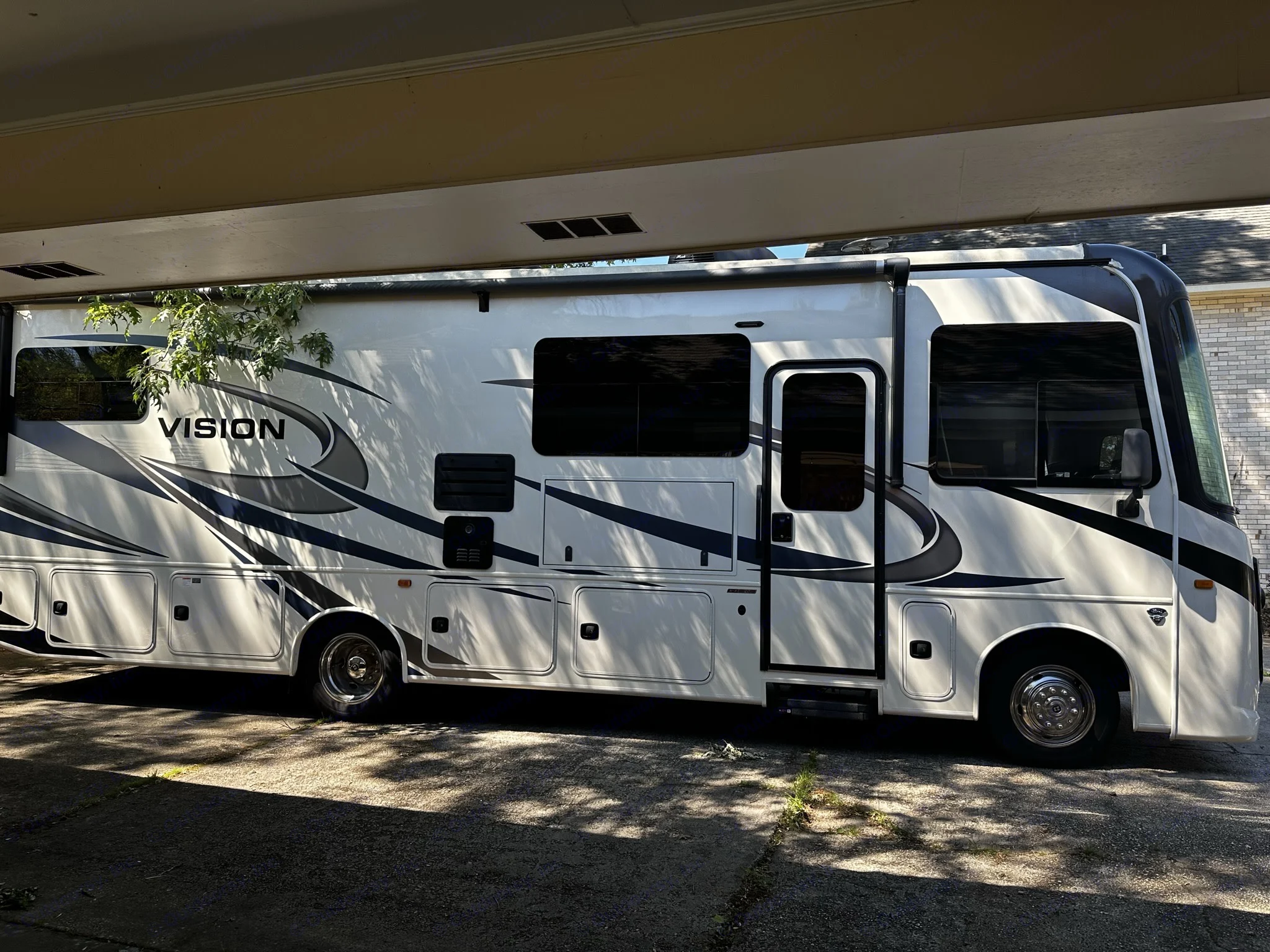 A large white and black RV, labeled 'VISION', is parked under a covered structure with dappled sunlight. Perfect for an rv rental experience.. Entegra Coach Vision 2021