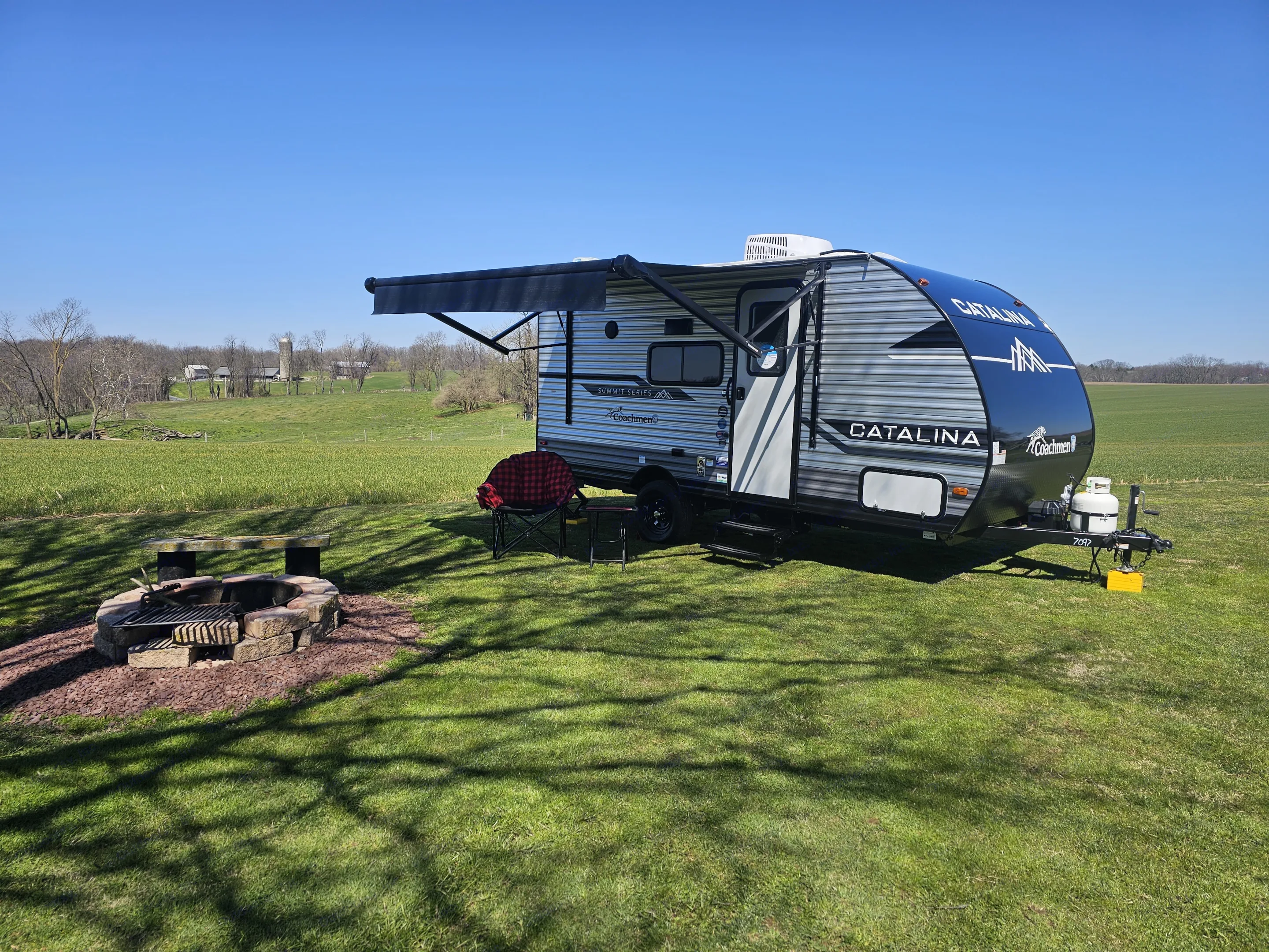 Scenic RV rental ready for adventure: travel trailer parked by fire pit on lush green grass under a clear blue sky.. Forest River Catalina 16BHX 2024
