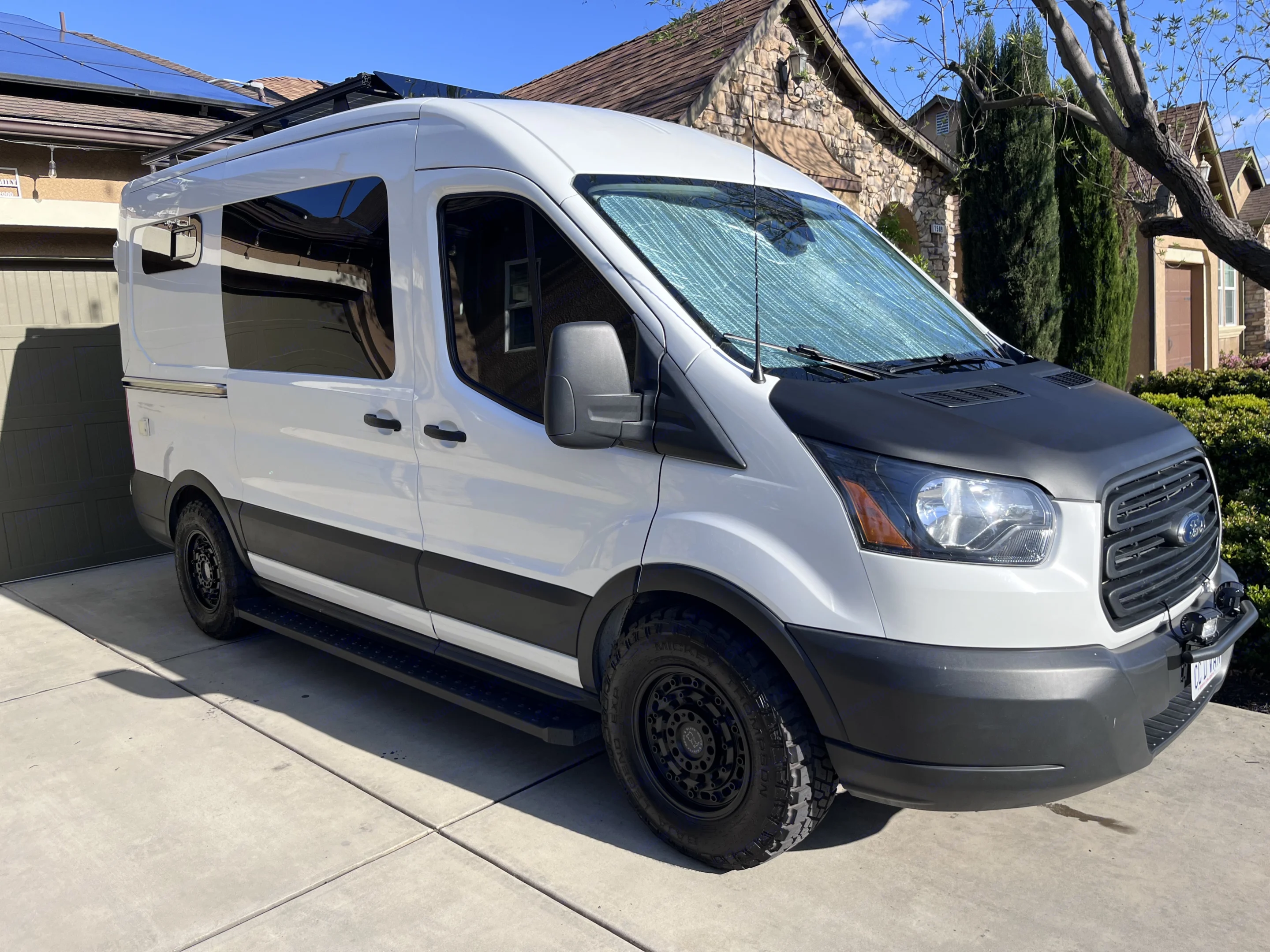 White Ford Transit camper van conversion, featuring solar panels, rugged tires, and a black hood. Perfect for an RV rental adventure.. Ford Transit 2019