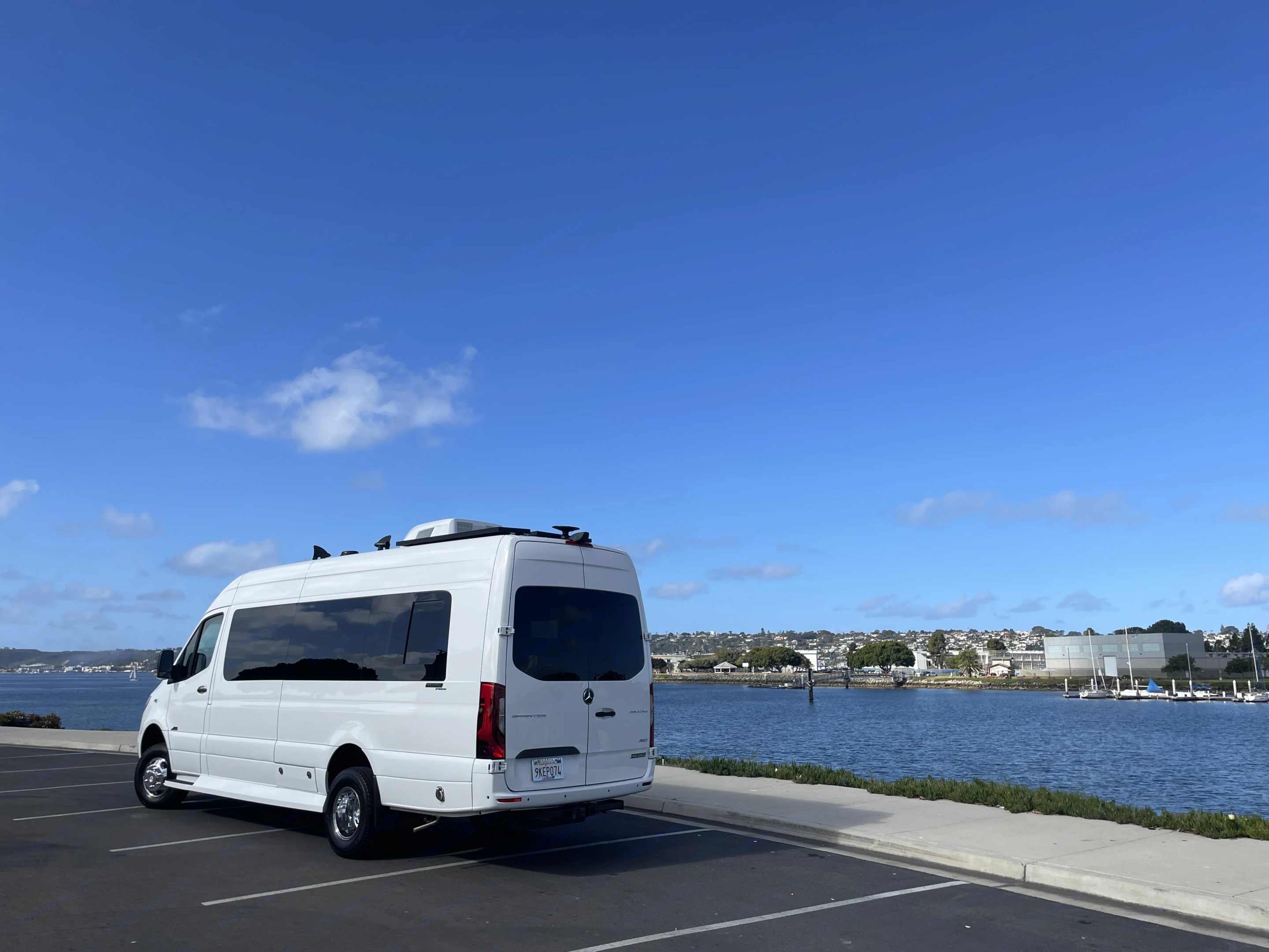 Explore the coast! White Mercedes Sprinter RV rental parked by a serene bay, with distant city and boats.. Mercedes-Benz Sprinter 2023