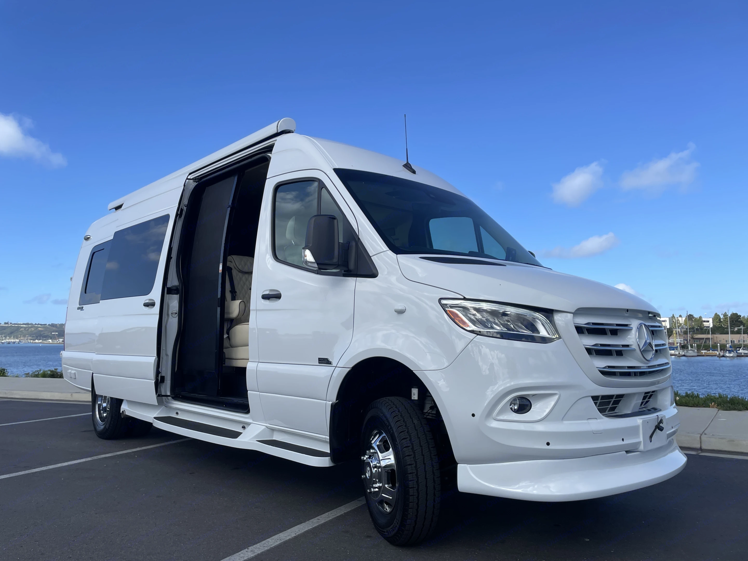 Luxury white Mercedes-Benz Sprinter RV rental, with its side door open, parked by a scenic bay with boats. Perfect for vacation!. Mercedes-Benz Sprinter 2023