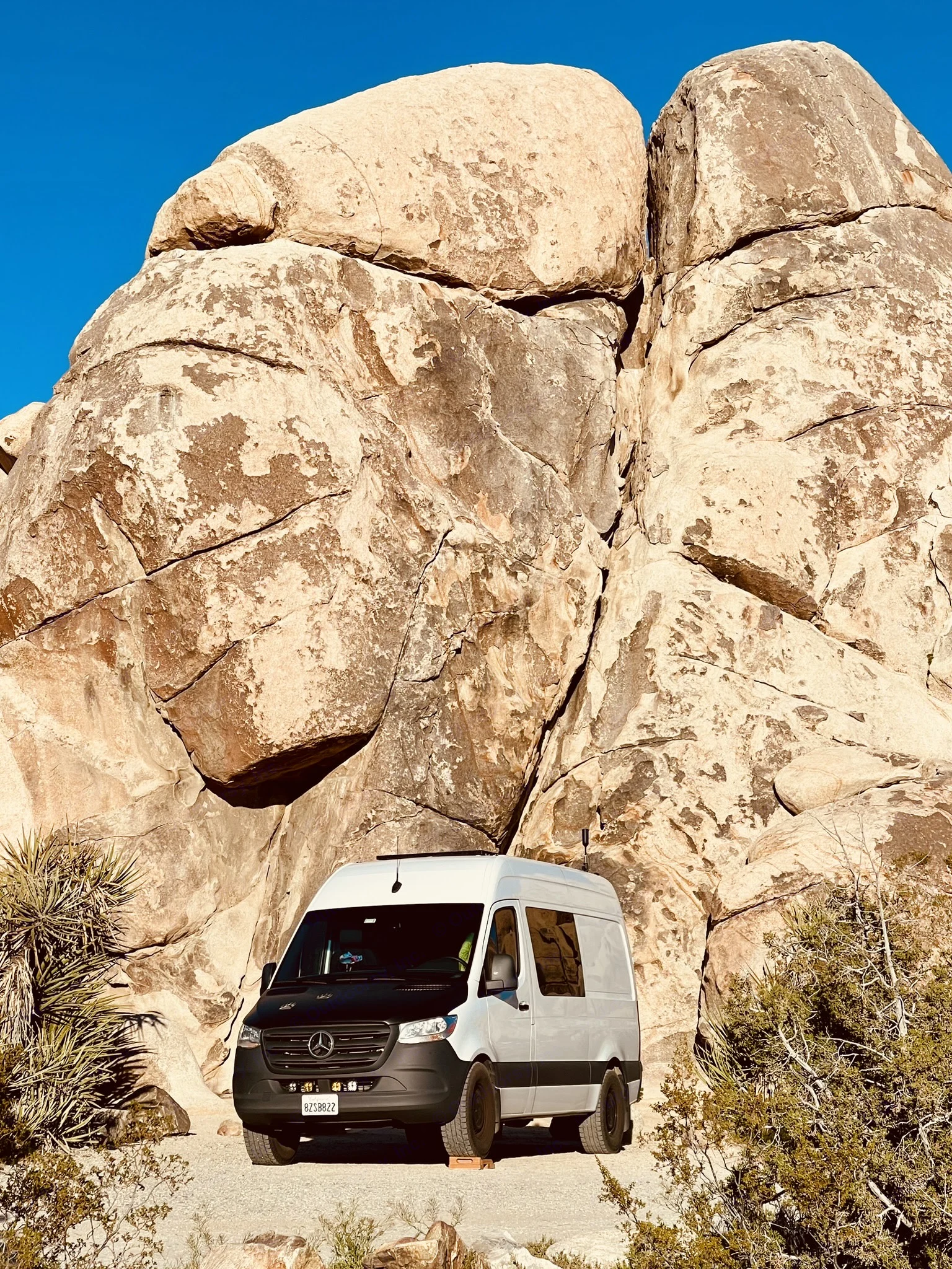 RV rental: White camper van parked by immense boulders and desert plants under clear blue sky, ideal for off-grid adventures.. Mercedes-Benz Sprinter 2021