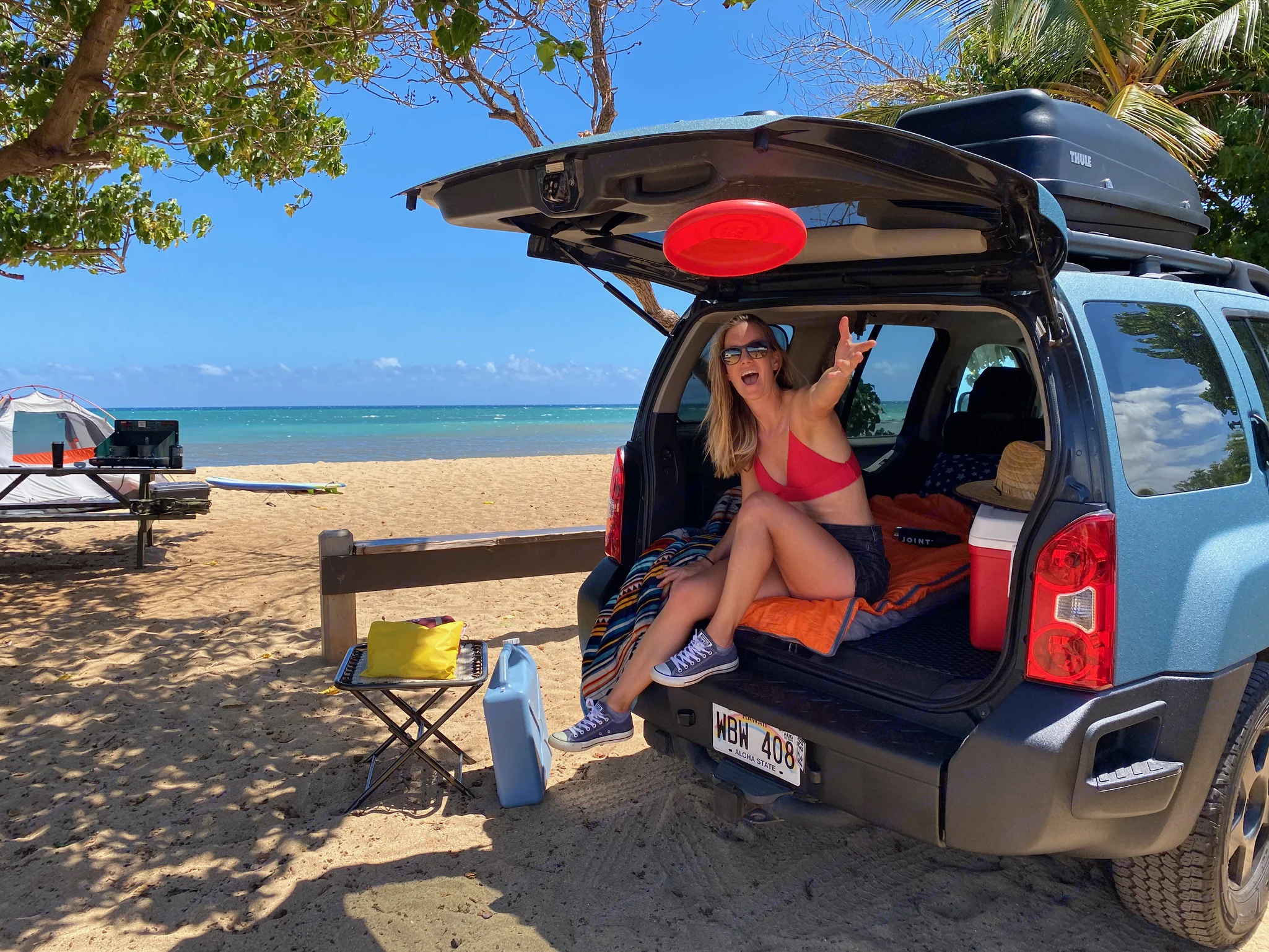 Smiling woman relaxes in the trunk of an SUV on a sandy beach, perfect for an adventurous trip like an RV. Nissan PRIMASTAR 2011
