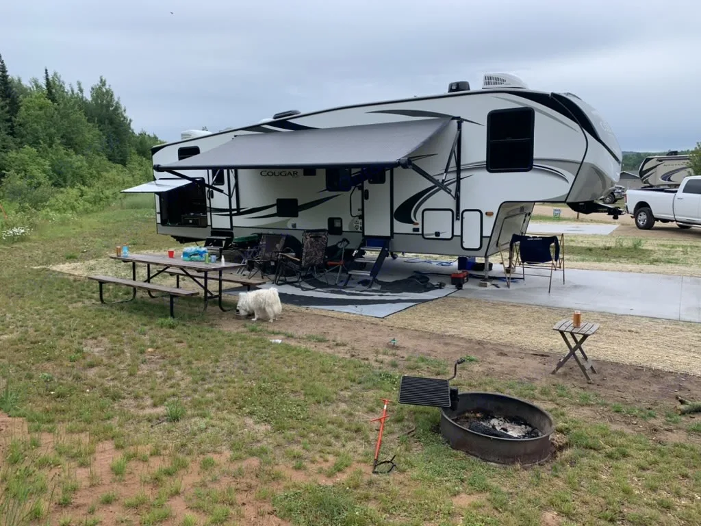 This spacious RV rental campsite features a white fifth-wheel, picnic table, fire pit, and a fluffy dog.. Keystone RV Cougar 2020