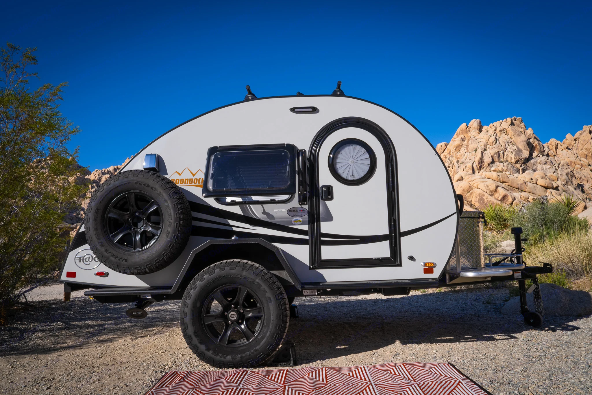 This compact teardrop RV rental, ideal for desert camping adventures, is parked on gravel with rocky terrain and a clear blue sky.. nuCamp T@G 2023