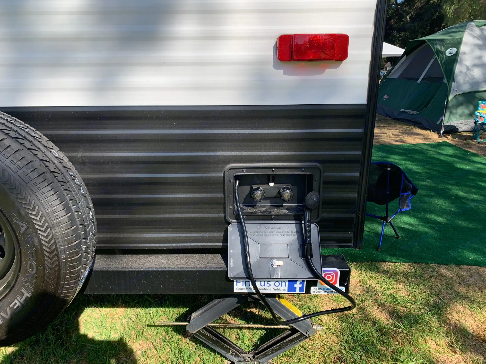 Back of an RV rental showing an outdoor shower utility panel, spare tire, and jack stand at a sunny campsite.. Clipper 18BHS 2023