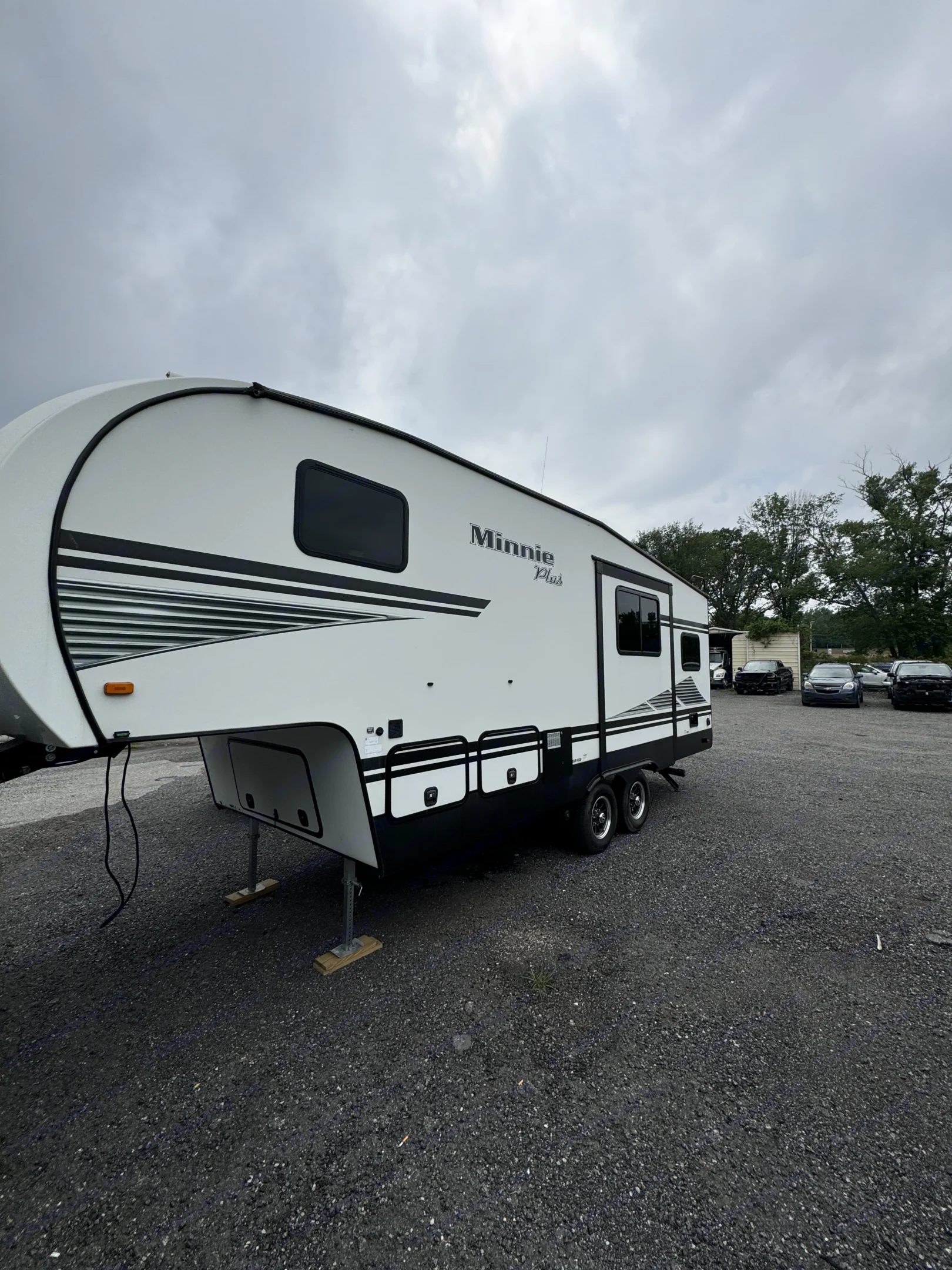 A large white Minnie Plus fifth-wheel RV, with black accents, is parked on a gravel lot under an overcast sky. Great for rv rental.. Minnie Minnie Plus 2019