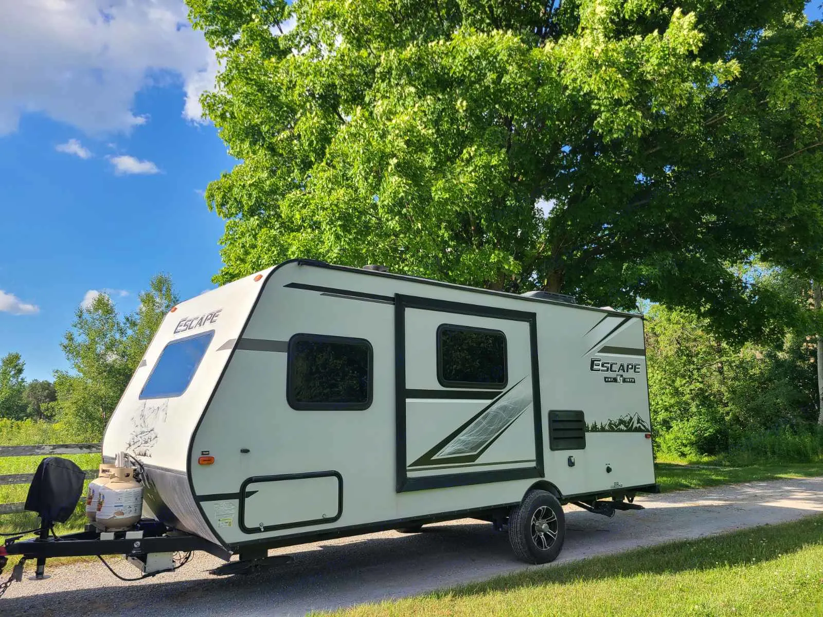 Light-colored Escape travel trailer parked on a gravel path amidst green trees & blue sky. Perfect for your next RV rental adventure.. KZ Escape 2020