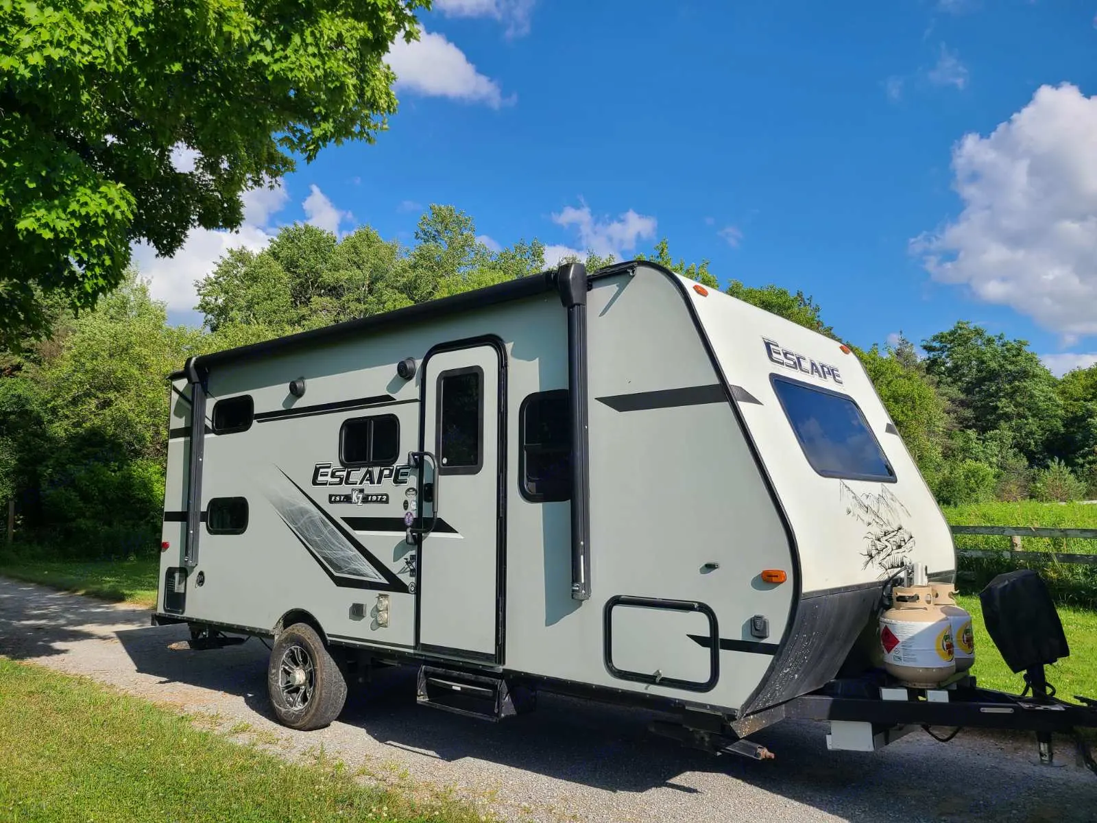 A light green Escape RV rental, a compact travel trailer, is parked on a gravel path amidst lush trees under a blue sky.. KZ Escape 2020