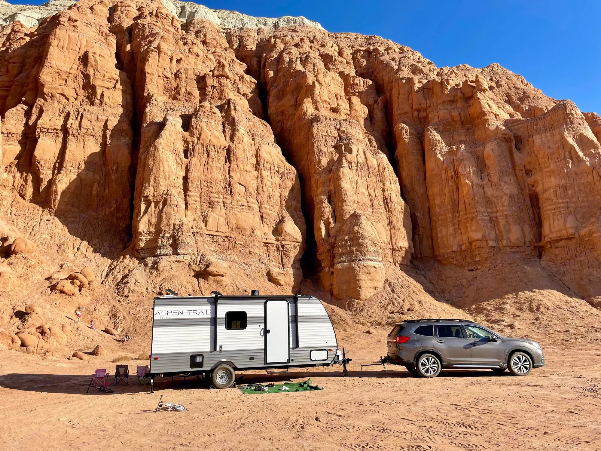 An RV rental and SUV parked amidst stunning red rock cliffs, with campers and kids exploring the sandy terrain. Perfect desert boondocking.. Dutchmen Aspen Trail 2024