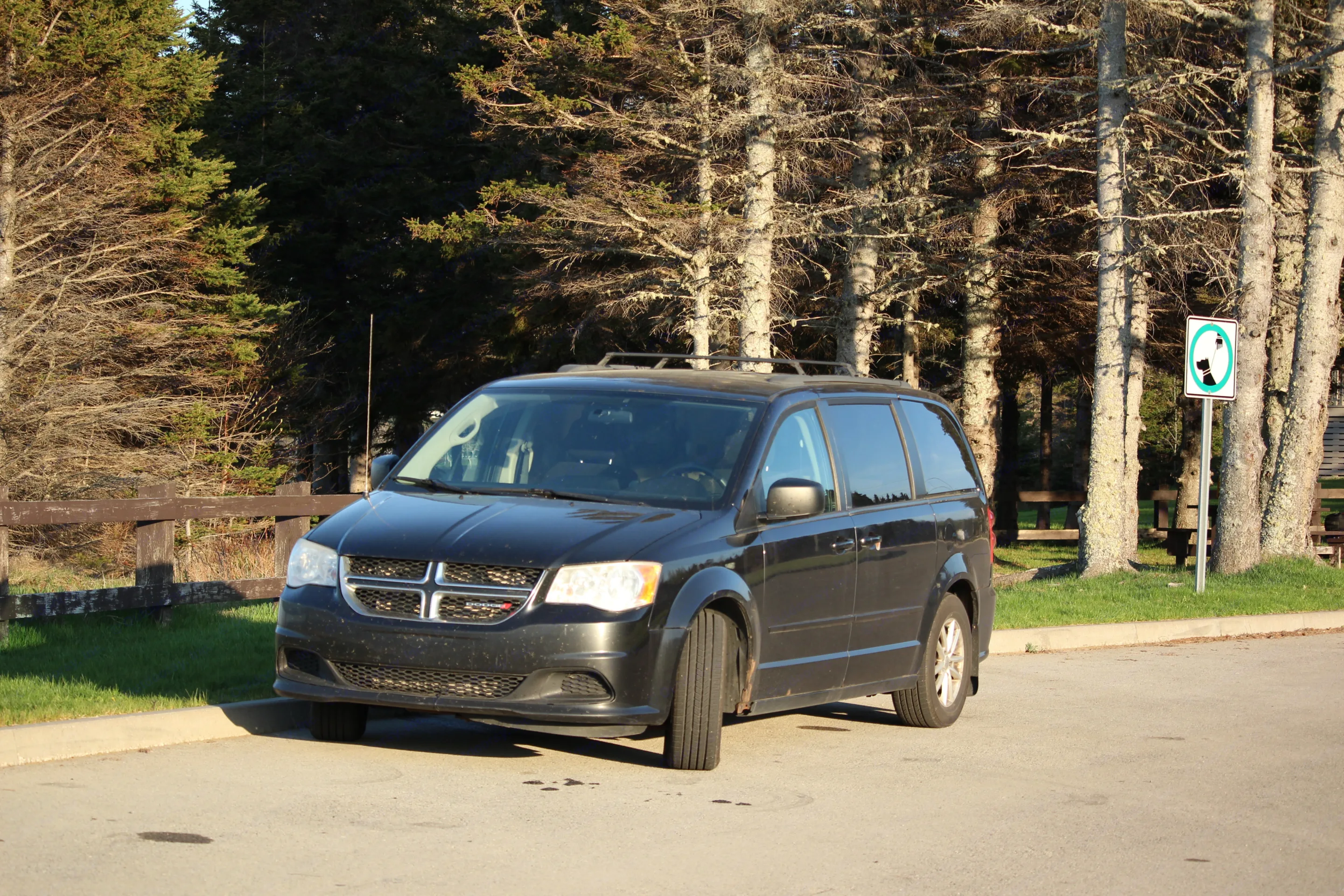 A black Dodge minivan on asphalt near trees & fence. Suited for travel; a small-scale RV rental alternative. 'No dogs' sign seen.. Dodge Grand Caravan 2013