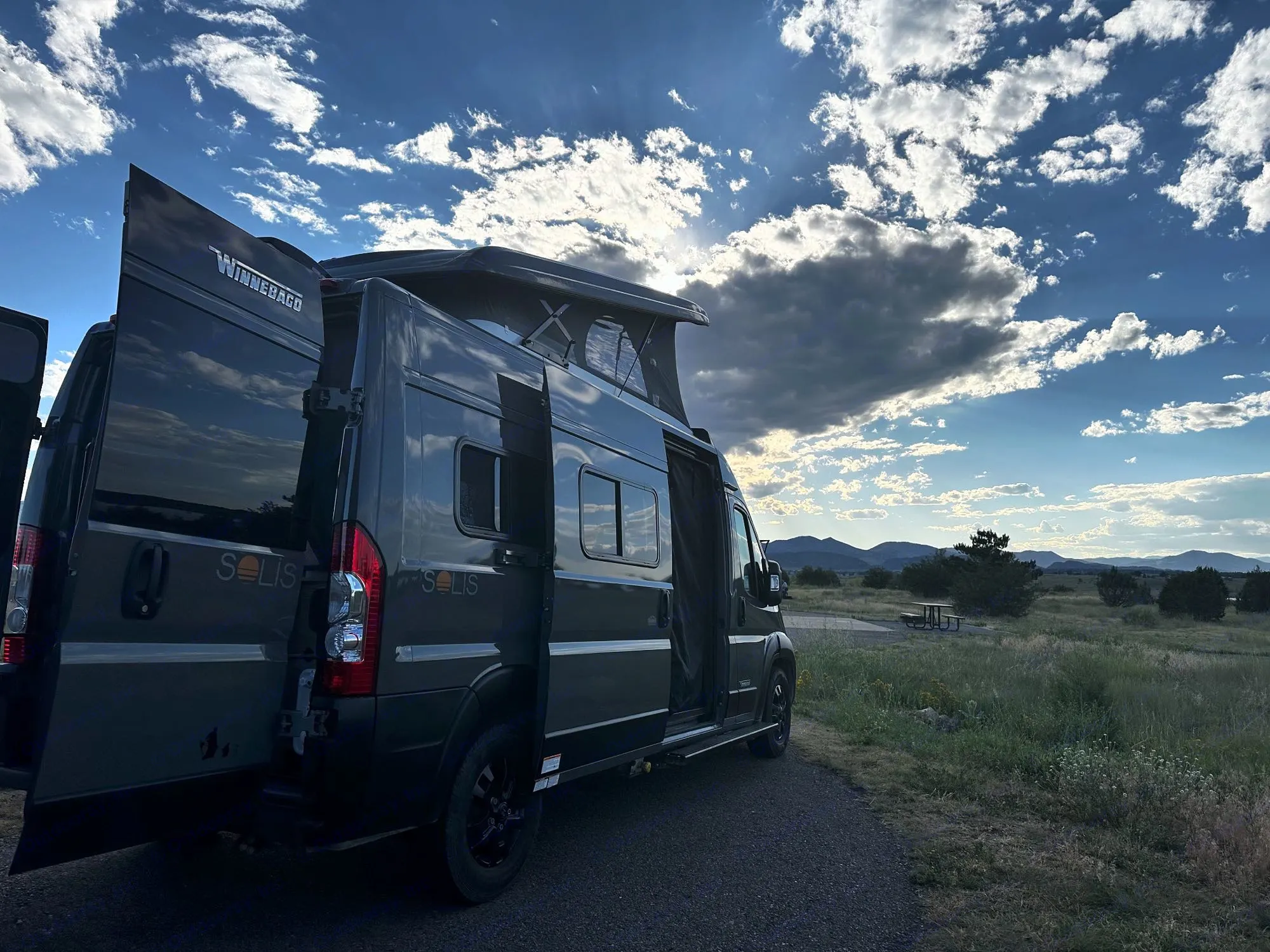 Dark gray Winnebago Solis rv rental van, pop-top up, doors open, in a scenic mountain landscape under a dramatic sky.. Winnebago Solis 2023