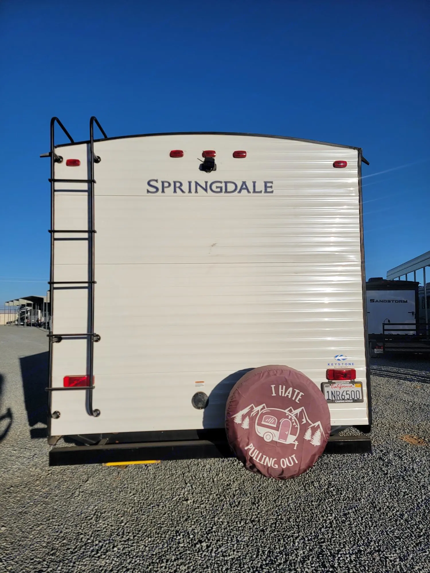 Rear view of a white Springdale travel trailer, possibly an RV rental, featuring a ladder and a humorous spare tire cover. Ready for camping!. Keystone RV Springdale 2021