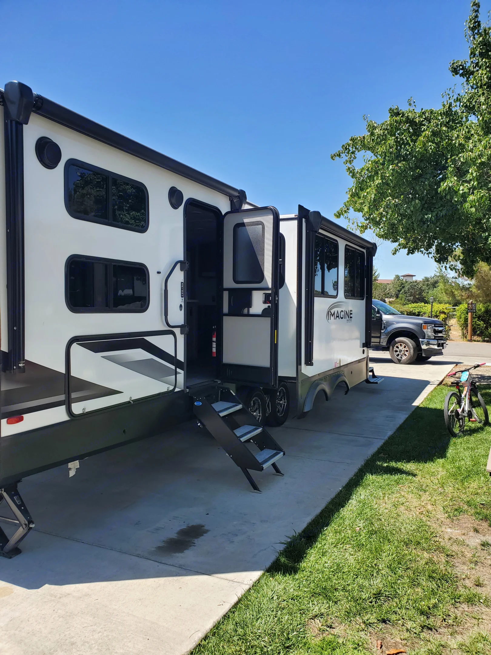 This Imagine RV rental is parked with open steps on concrete, ready for a camping trip. A pickup truck & bike are nearby under blue sky. Grand Design Imagine 2023