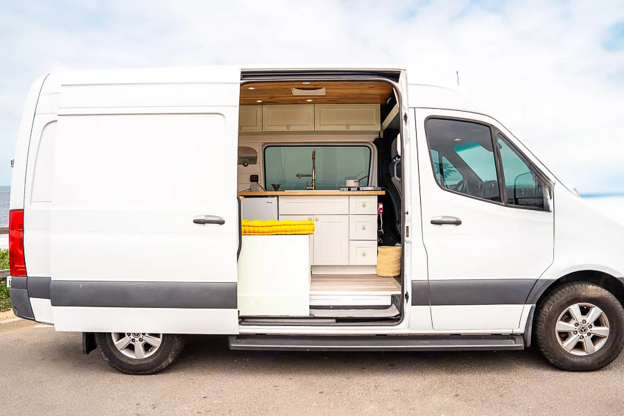 This custom white camper van, perfect for an RV rental, shows its open side door revealing a stylish kitchen and cozy interior setup for travel.. Mercedes-Benz Sprinter 2019